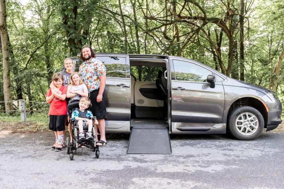 Family stands near a gray accessible van with ramp deployed in a wooded area.