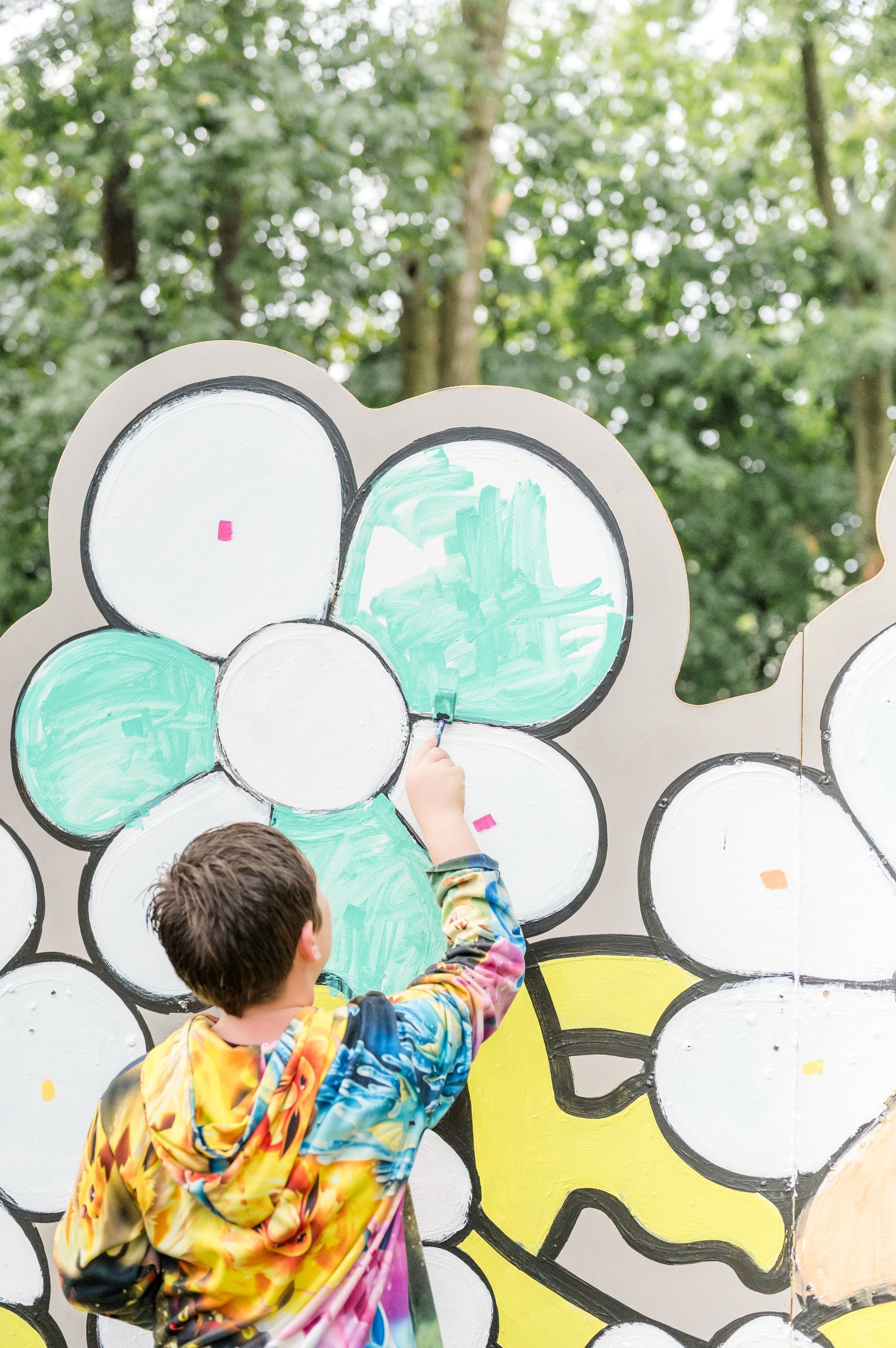 Person in colorful hoodie painting green flower on a mural outdoors.