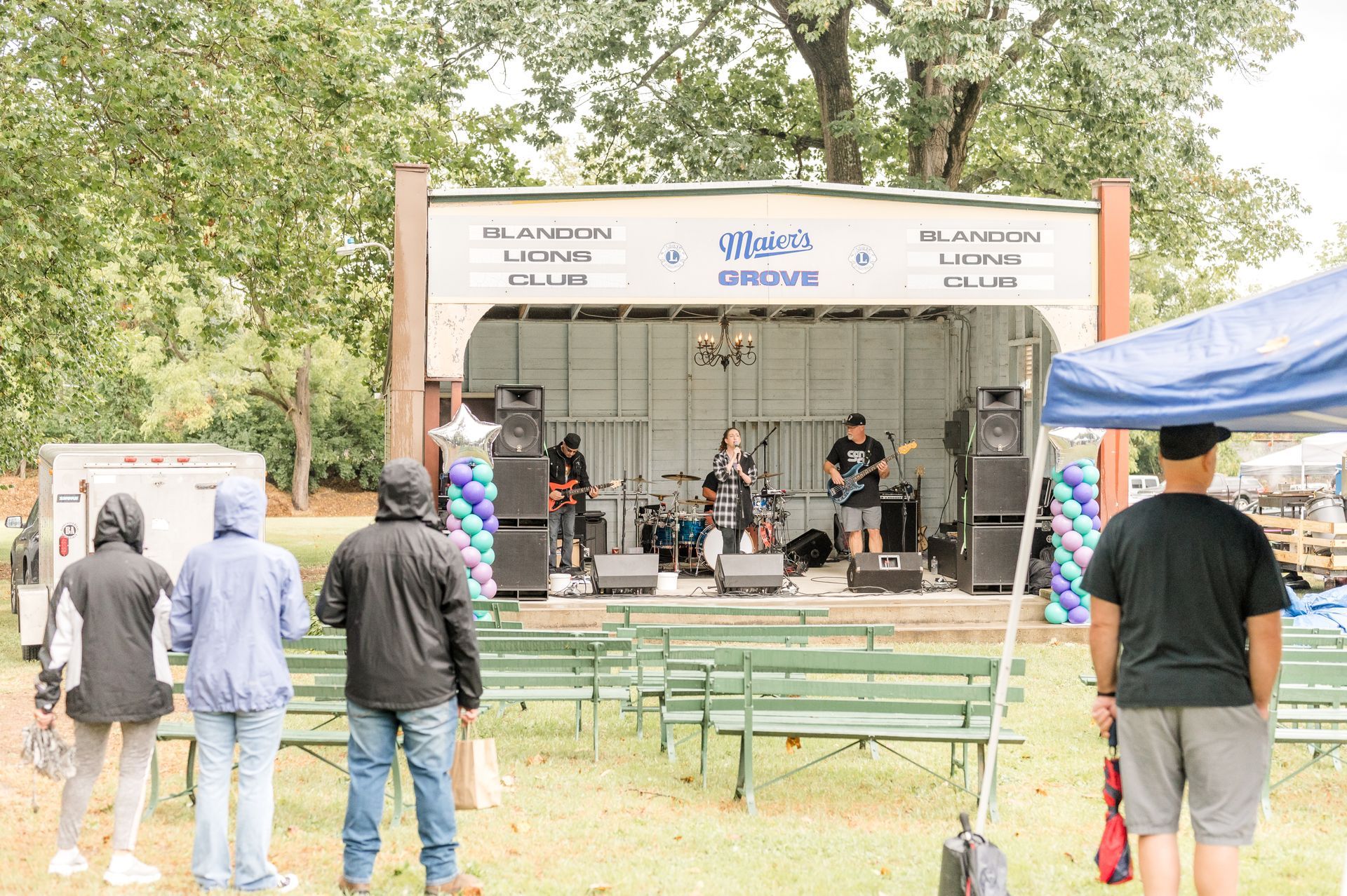 Band performing on outdoor stage; people watching in front. Gray sky, green trees.