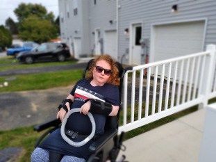 Person in a wheelchair with arm brace and sunglasses outdoors near a white ramp and building.