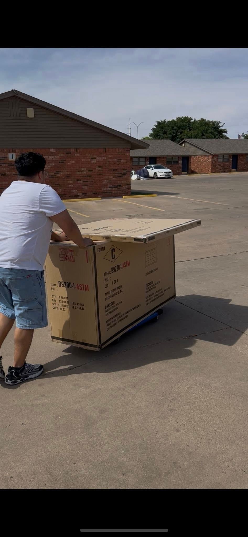 Person pushing large cardboard box on a dolly outside brick buildings under a sunny sky.