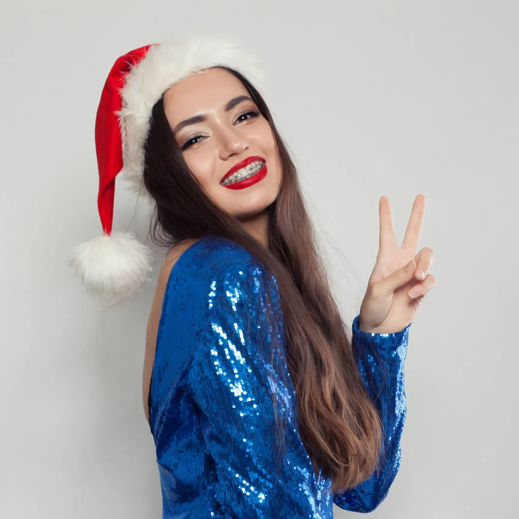 Woman with braces, wearing a Santa hat and a blue dress, smiling and giving the peace sign