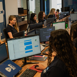 Pessoas trabalhando em computadores em um ambiente de escritório. Elas estão sentadas em uma mesa comprida com monitores e notebooks.