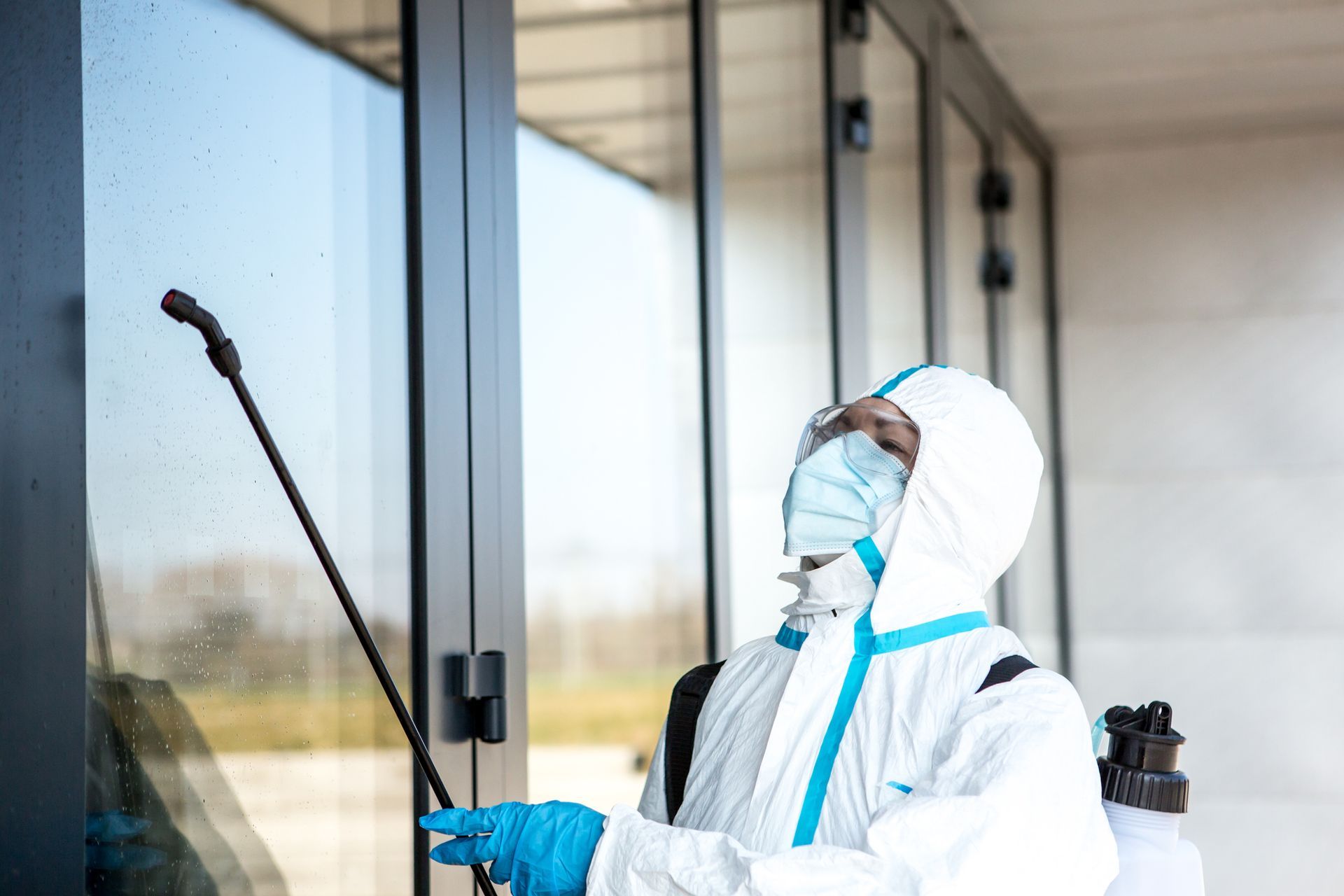 Person in protective suit disinfecting glass doors with spray wand.