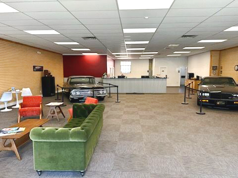 Exterior of a grey building with a Cox sign, a loading bay, three green trash bins, and a blue car parked on the side.