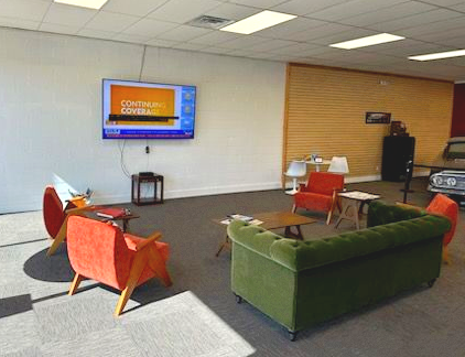 Exterior of a grey building with a Cox sign, a loading bay, three green trash bins, and a blue car parked on the side.