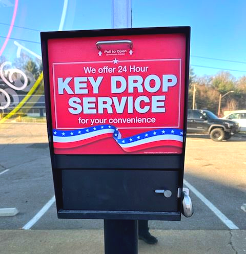 Exterior of a grey building with a Cox sign, a loading bay, three green trash bins, and a blue car parked on the side.