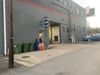 Exterior of a grey building with a Cox sign, a loading bay, three green trash bins, and a blue car parked on the side.