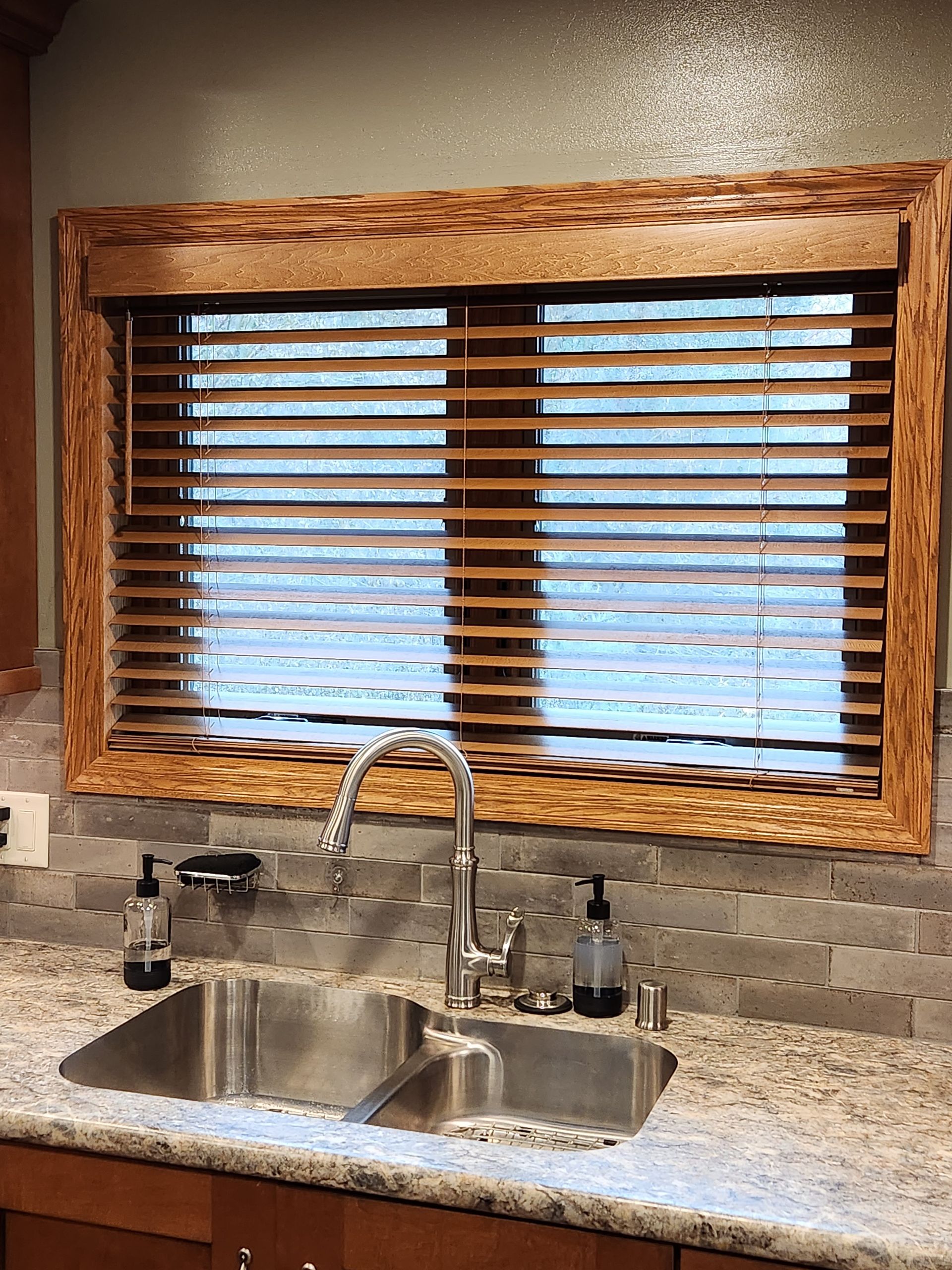Kitchen sink under a window with wooden blinds and frame. Granite countertop and tiled backsplash.