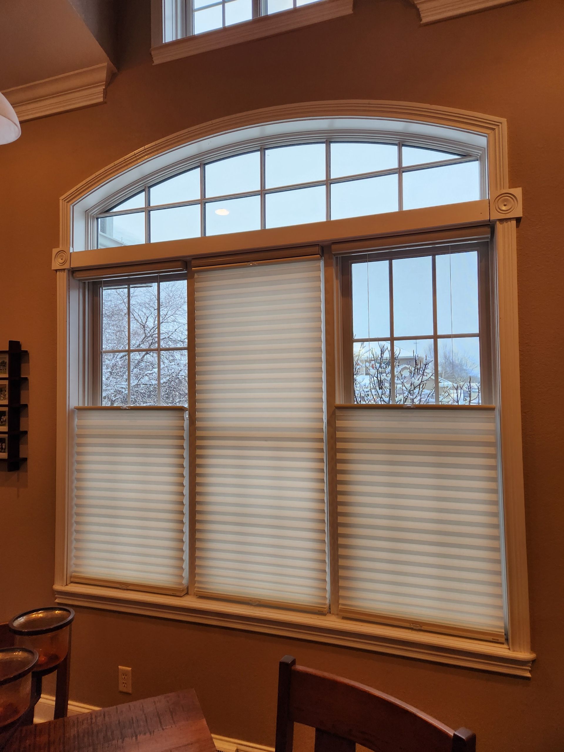 Brown-walled room with a large window. Tan blinds cover the lower panes, and the upper arched window has no covering.