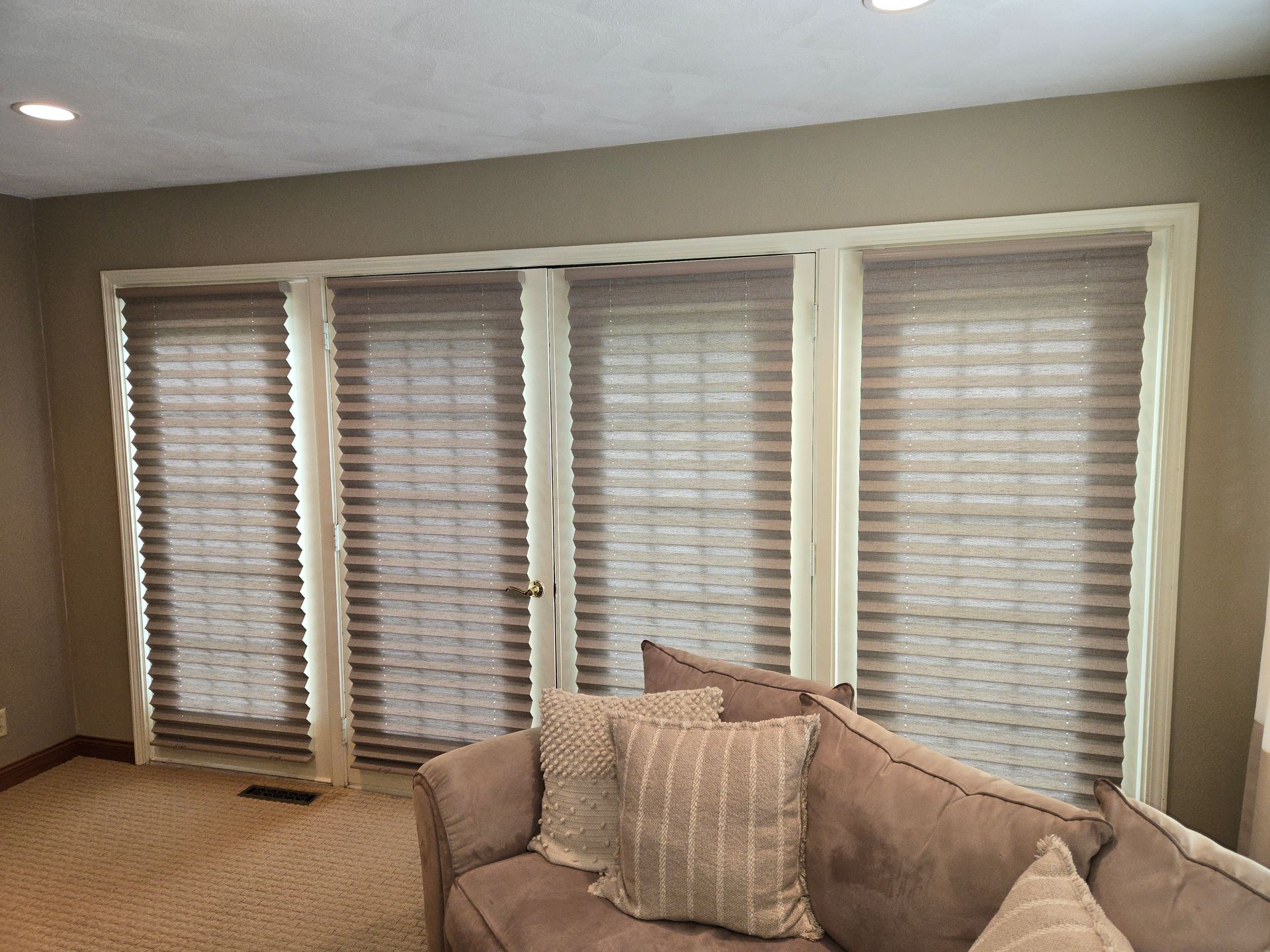 Living room with beige walls, windows with pleated shades, and a brown sofa with decorative pillows.