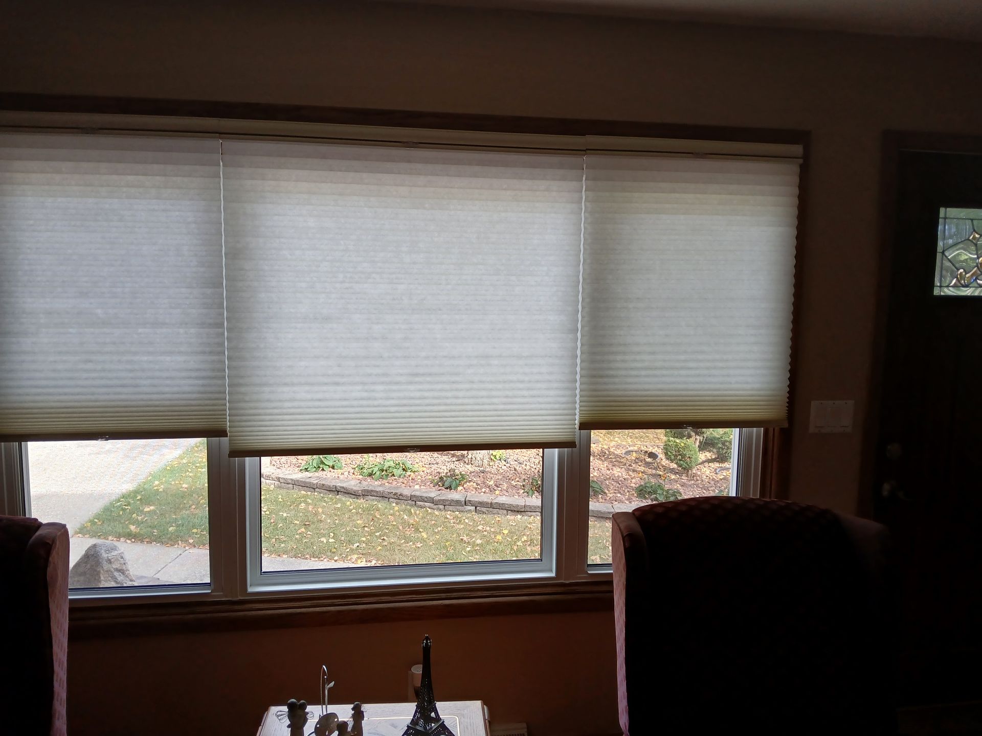 Three light-colored pleated blinds cover a window overlooking a yard. A dark door is on the right.