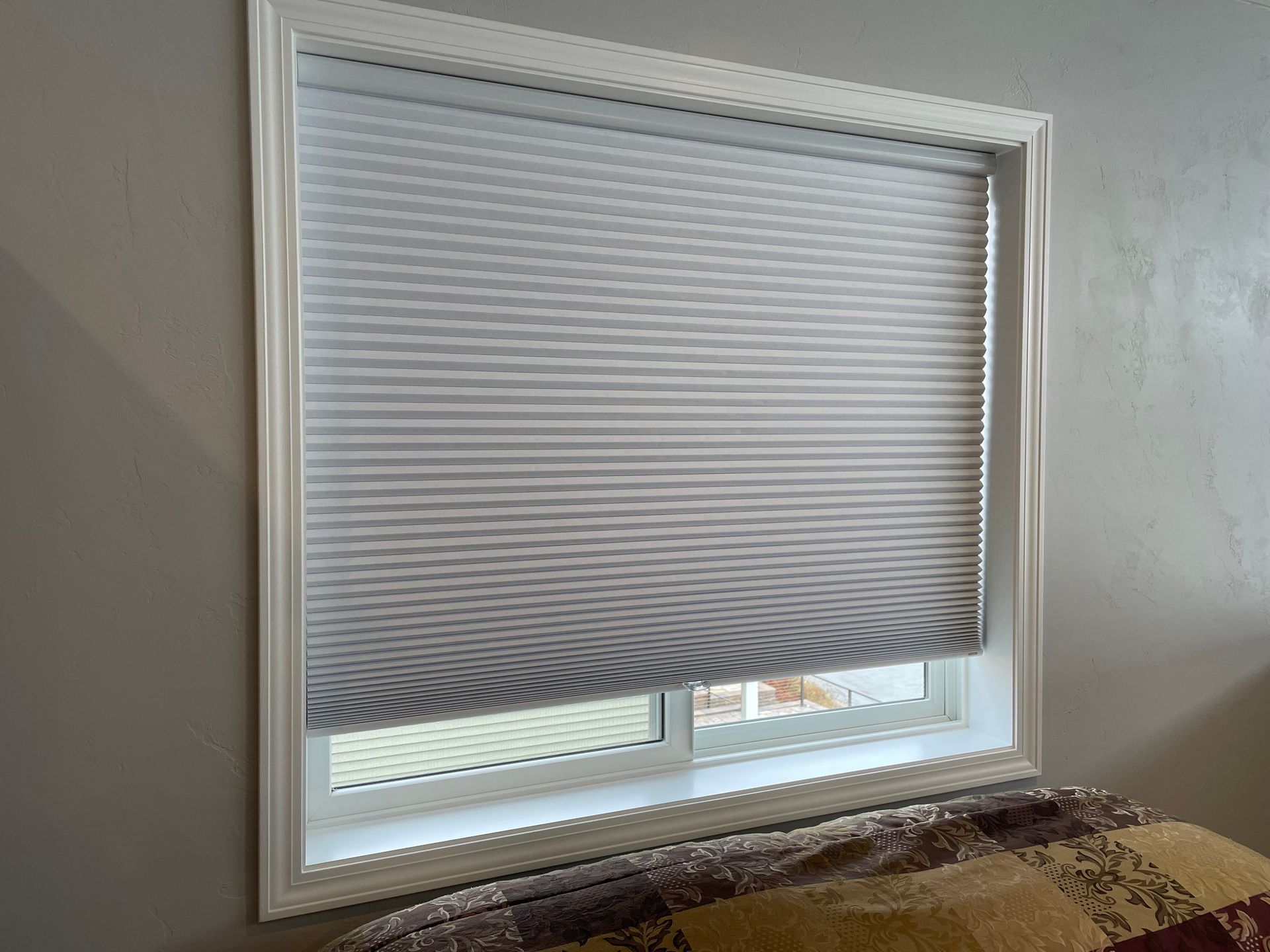Window with gray pleated shade, white frame, against a gray wall. Partially visible bed below.