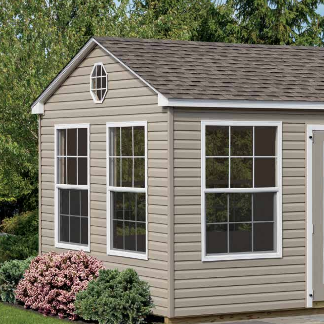 Tan shed with three windows, oval window, and shingled roof, set against trees.