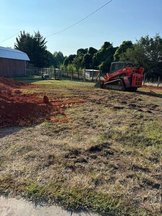 A red skid steer operates on a grassy construction site next to a fence and barn, under a clear blue sky.