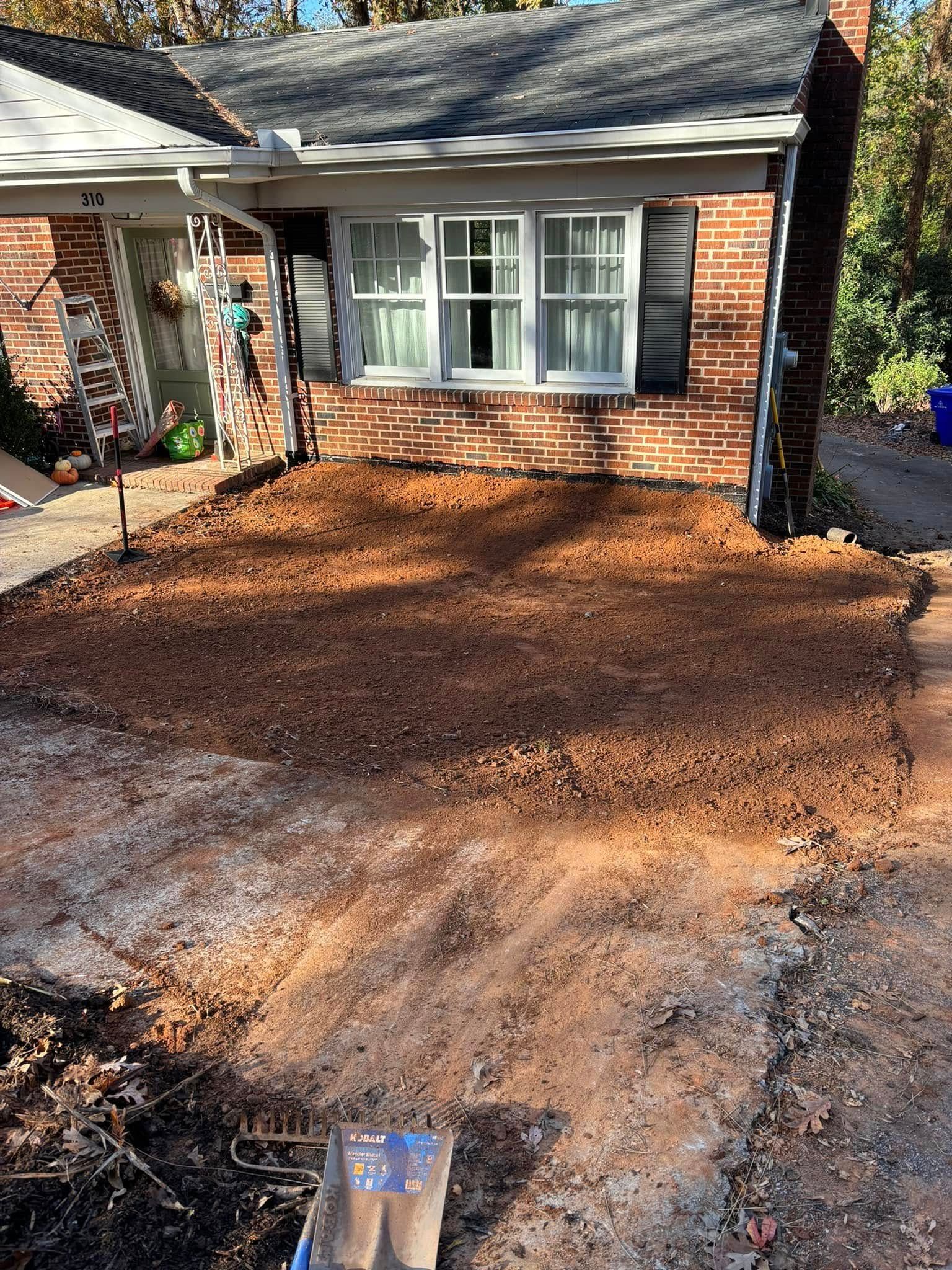 A front yard with freshly turned soil in front of a brick house with a window and black shutters.
