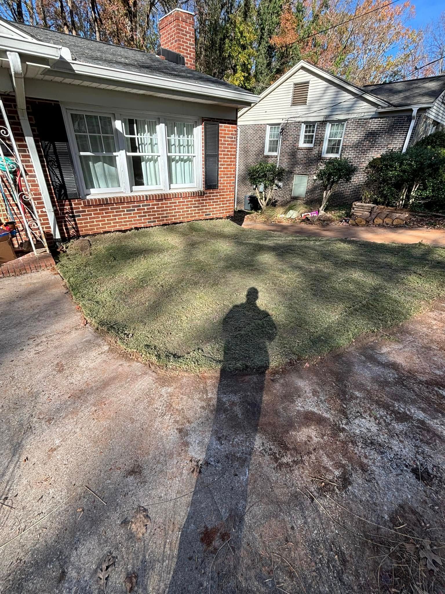 Shadow of a person standing in a yard in front of a brick house with a garden. The yard is grassy and the ground is gravel.