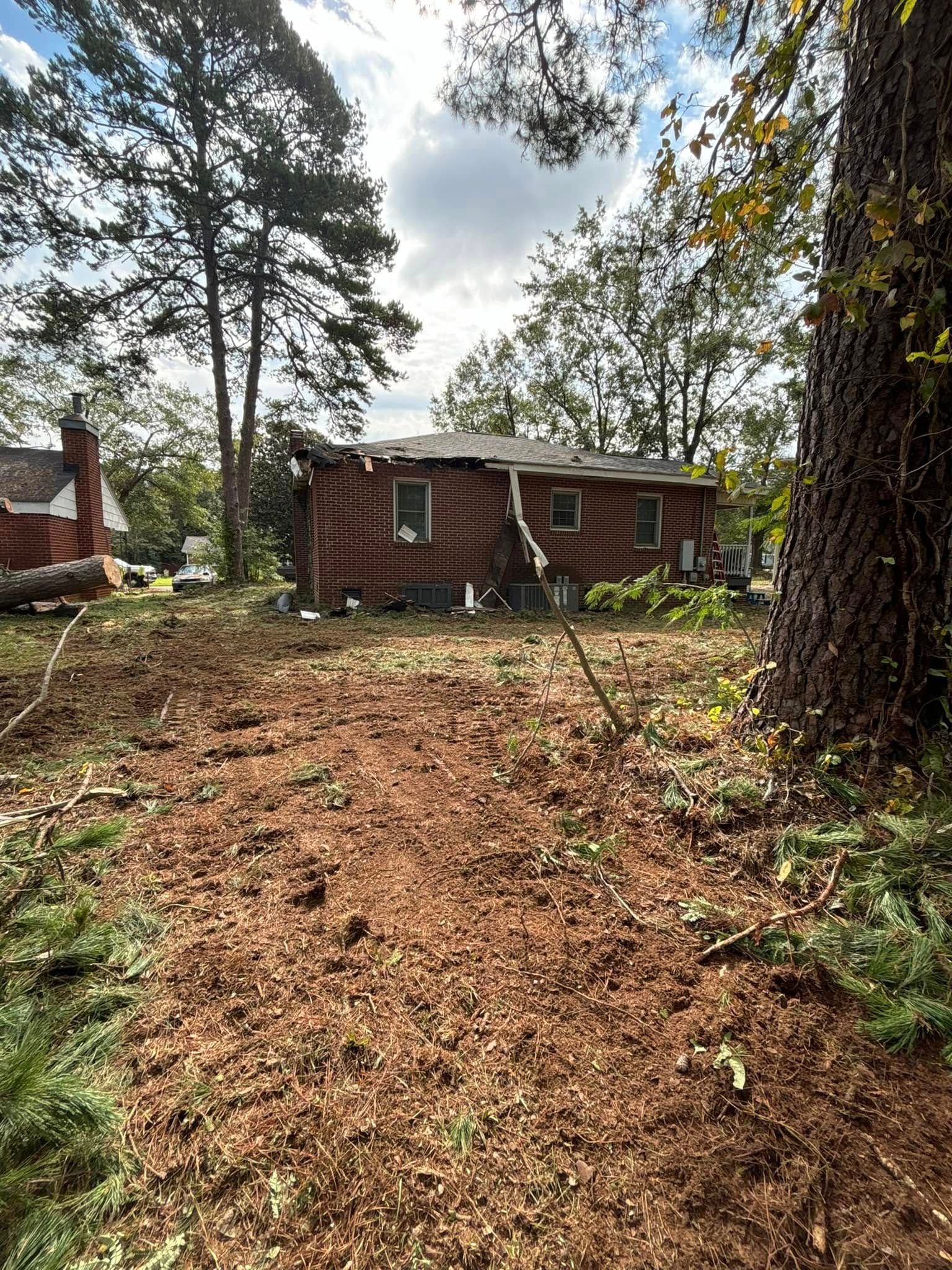 A damaged, single-story house with a collapsed wall amidst a clearing, surrounded by trees under a cloudy sky.