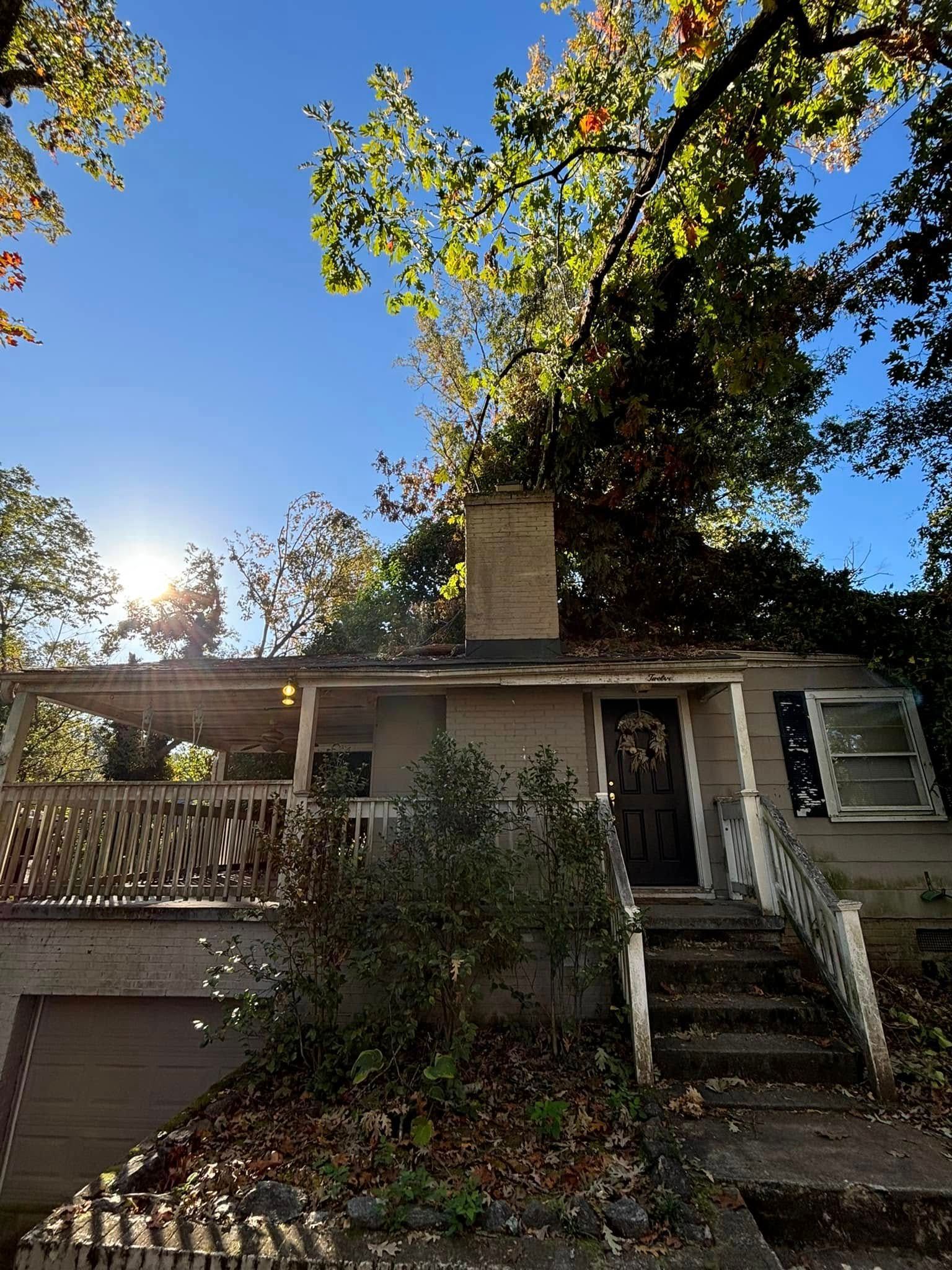 A small, beige house with a porch and chimney surrounded by trees under a bright blue sky. Sunlight shines from the left.