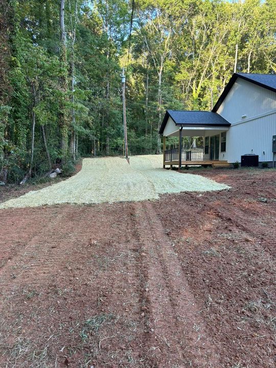 Dirt path leading to a light-colored gravel area next to a house with a porch in a wooded setting.