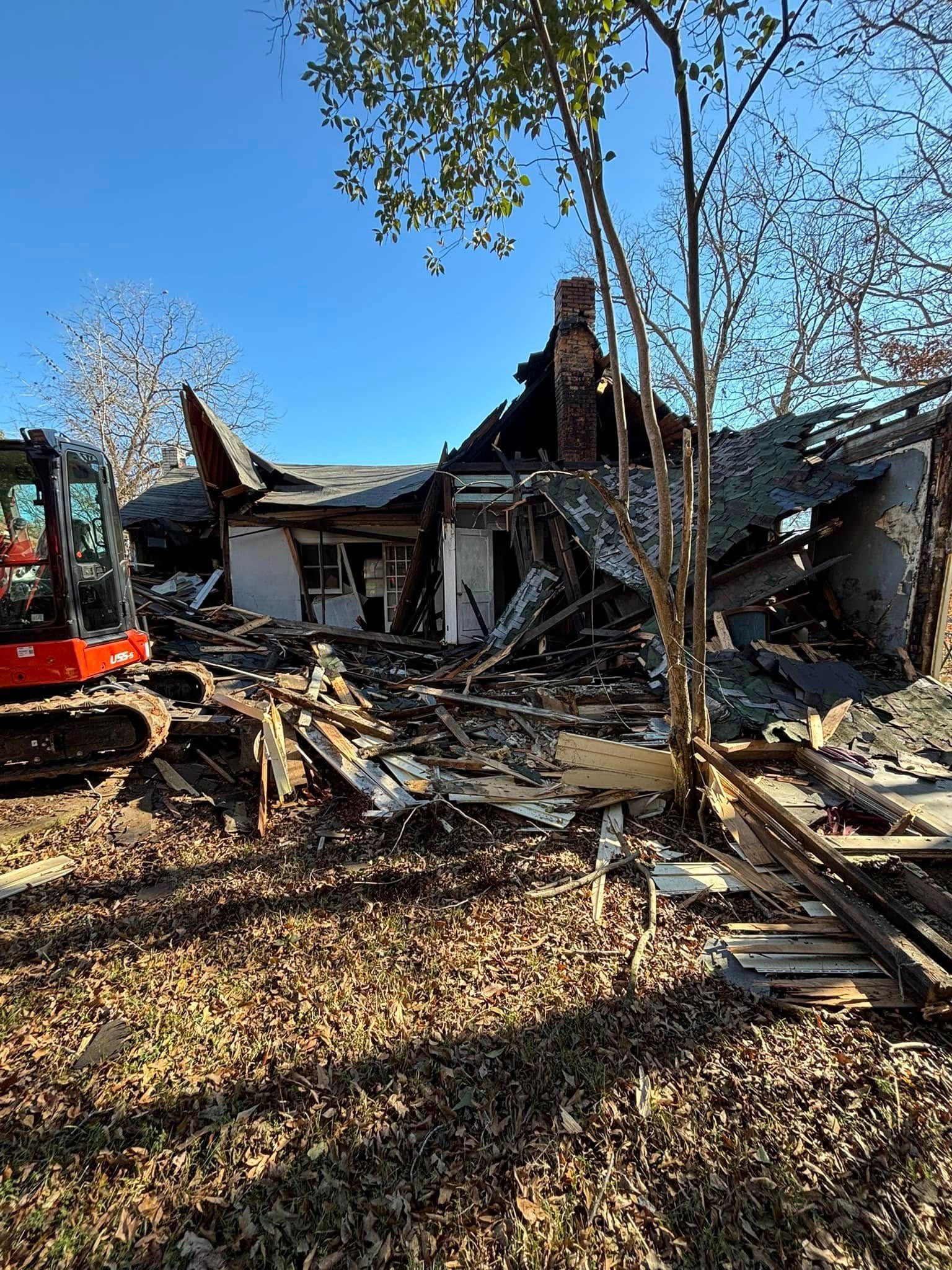 A demolished house sits among scattered debris, with a small excavator in the left foreground under a clear blue sky.