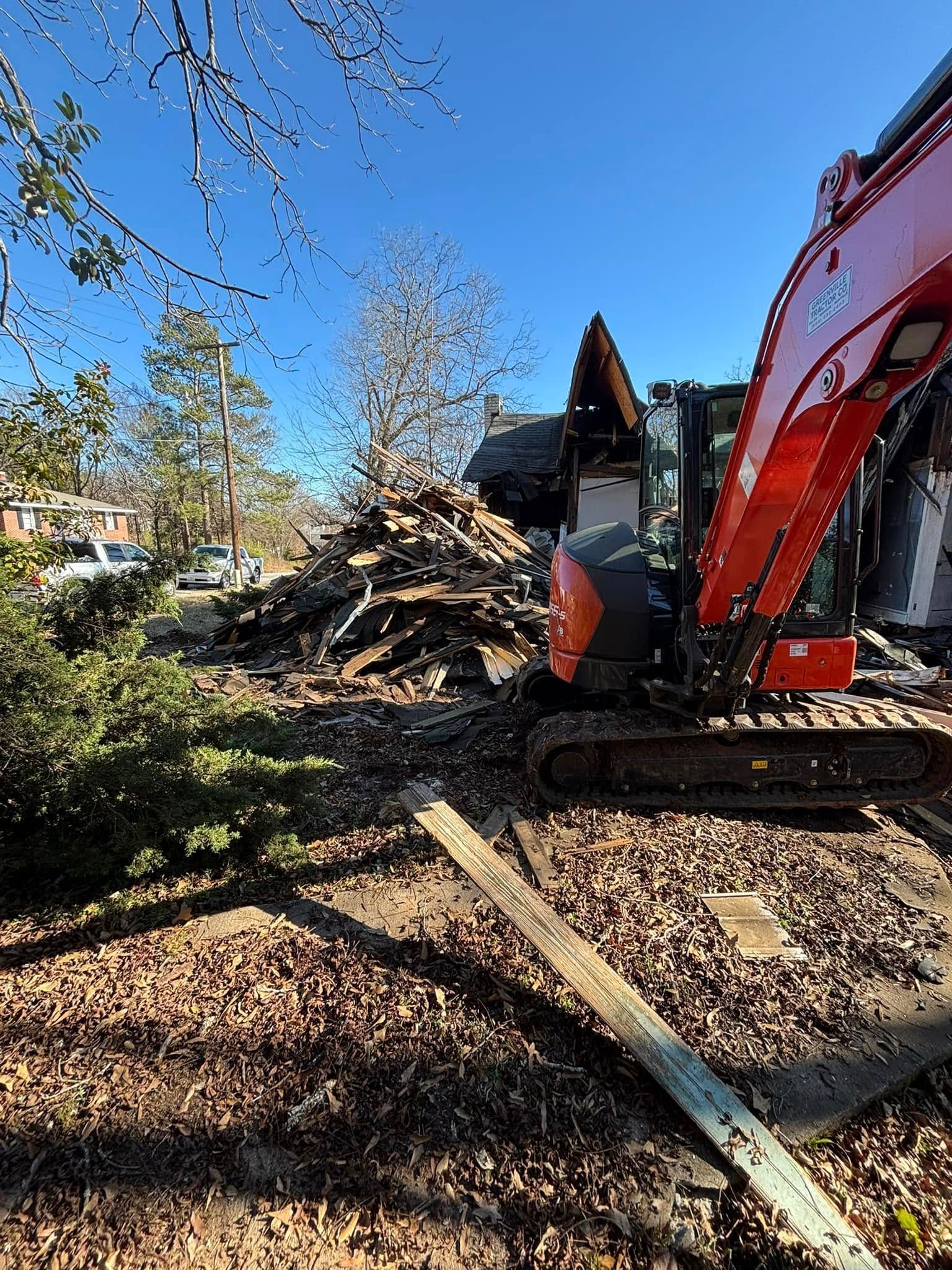 A red excavator demolishes a structure, with debris and wood scattered on the ground under a clear blue sky.