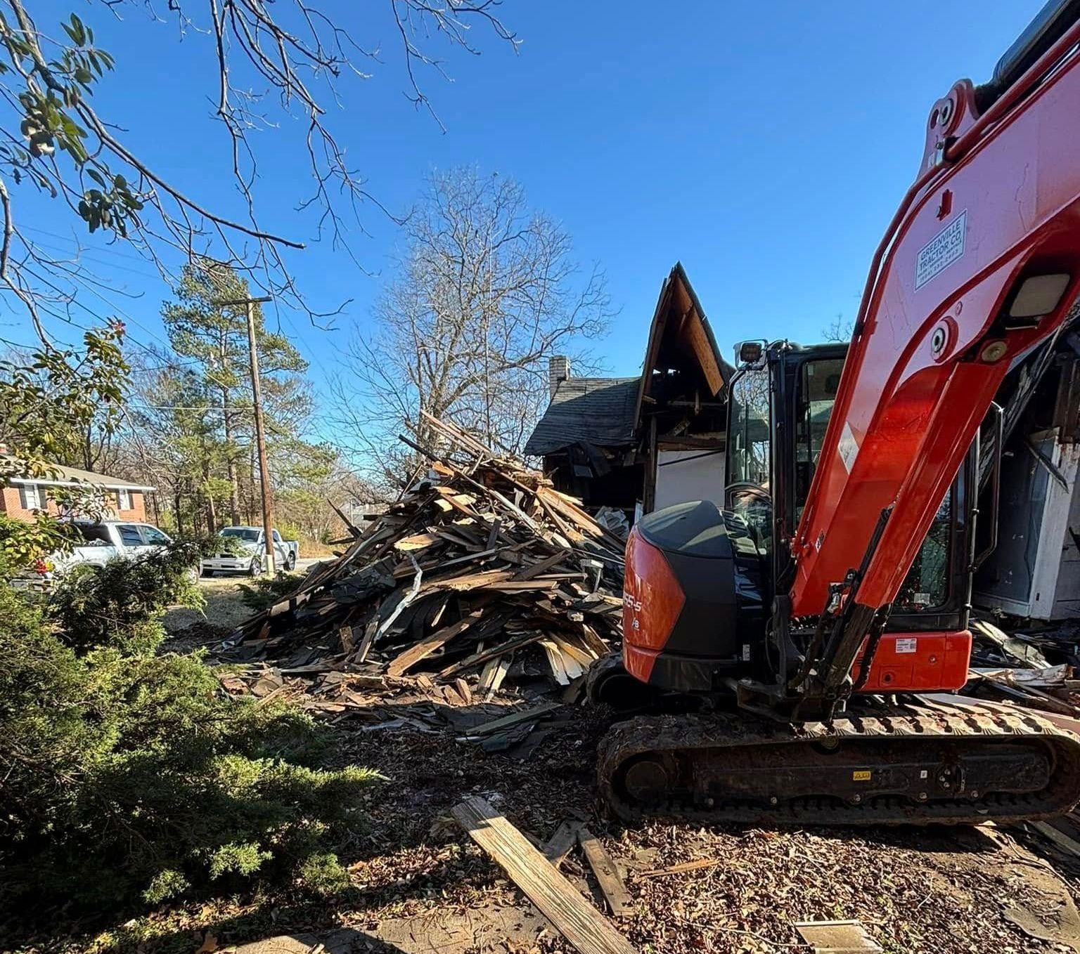 A red excavator demolishes a building on a sunny day, surrounded by debris and fallen leaves.