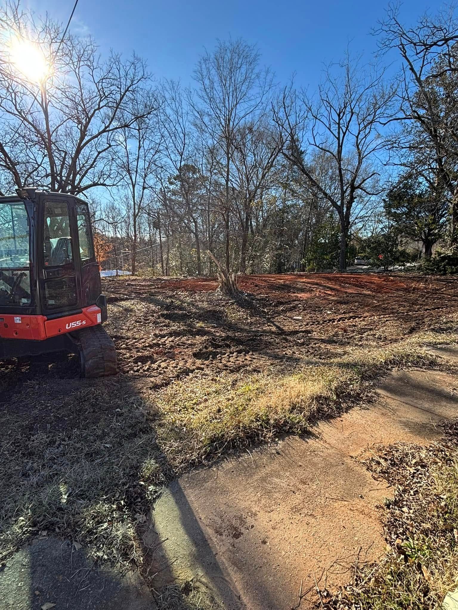 A small construction vehicle on a dirt lot, clearing debris, trees in the background, bright sunlight.