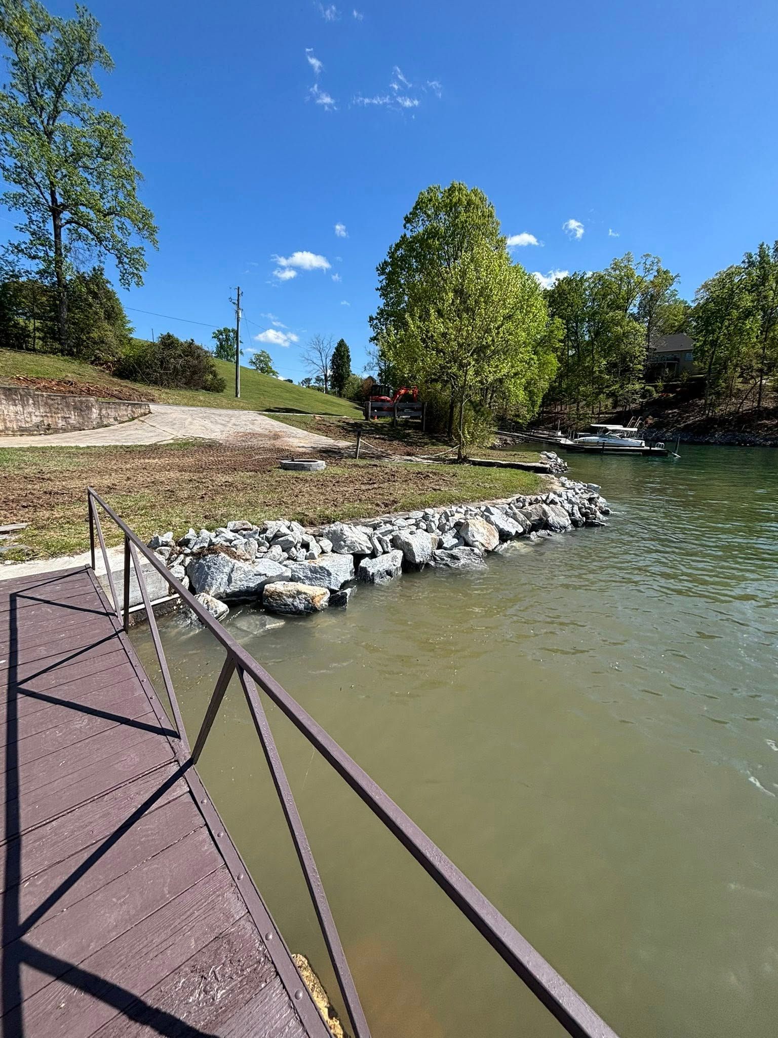 View from a dock overlooking a rocky shoreline, trees, and a lake under a blue sky. A metal railing is in the foreground.