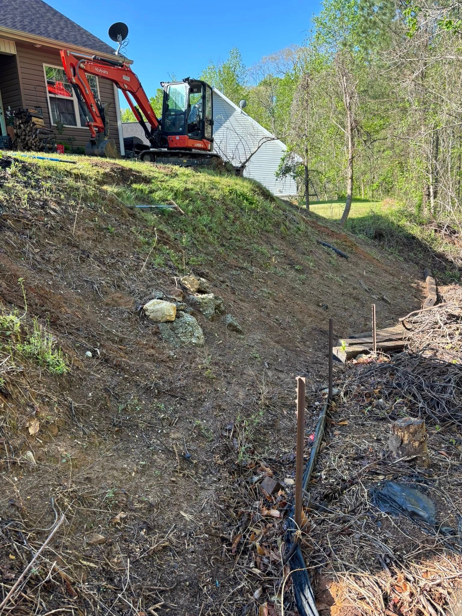 An excavator on a hillside clearing debris near a house. The ground is covered in mulch and old retaining wall remnants.