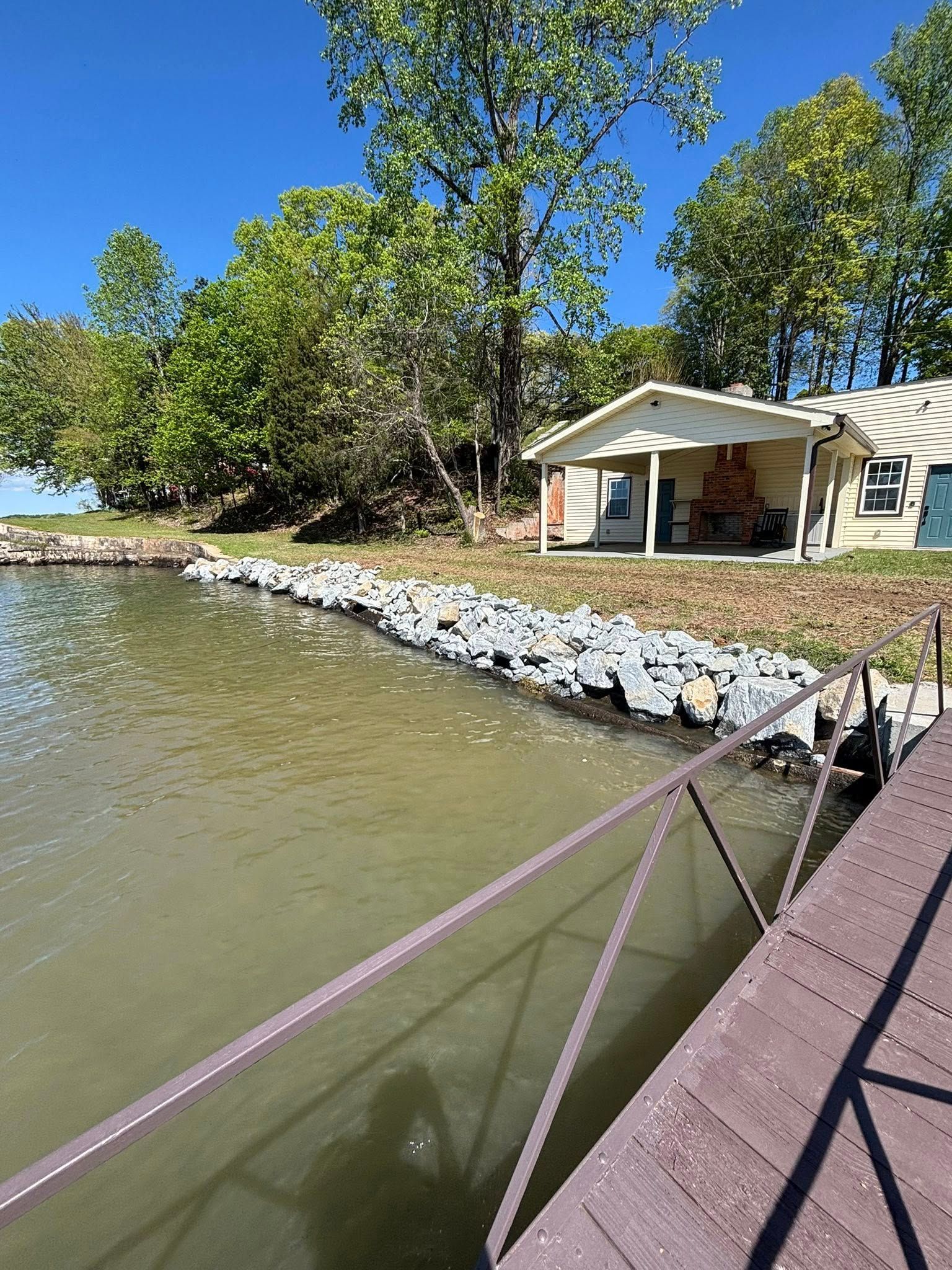 A waterfront property with a rock-lined bank, dock, and small beige house with trees in the background under a blue sky.