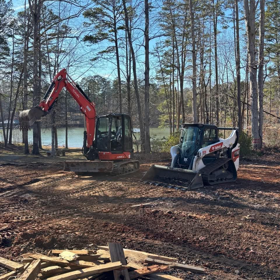An excavator and Bobcat on a dirt lot, clearing land near trees and a body of water. Sunny day.