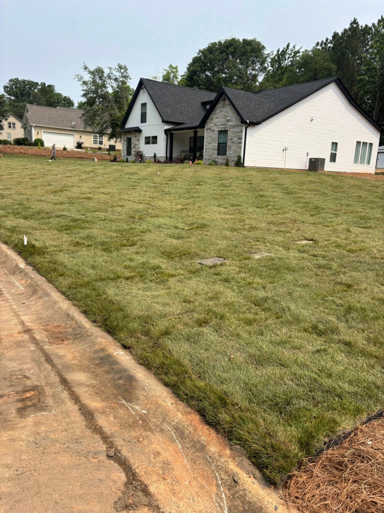 A newly built house with a dark roof and light siding sits on a large, grassy lawn next to a concrete curb.