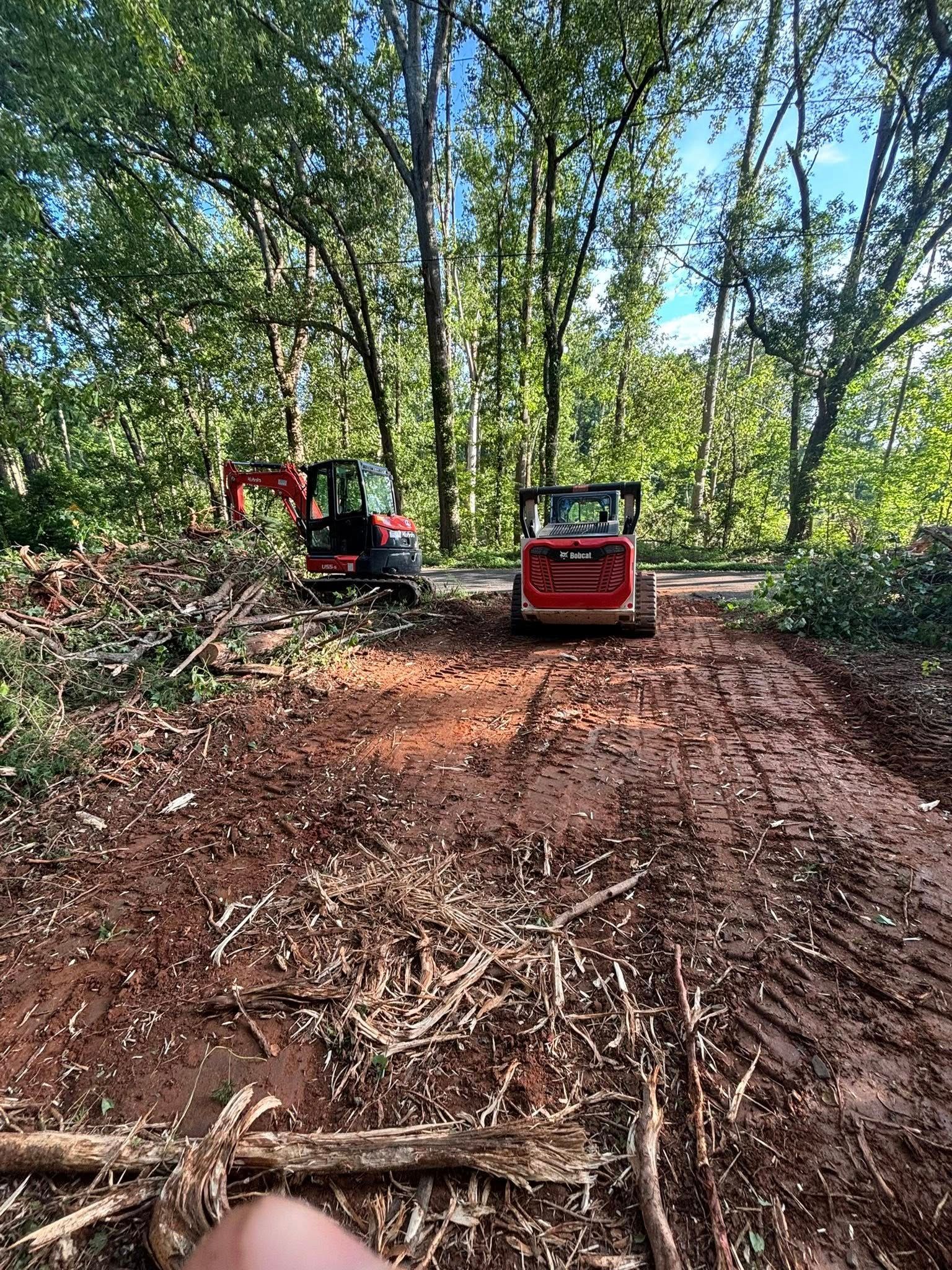 An excavator and skid steer on a muddy path in a wooded area after clearing brush. Red machines, brown soil, green trees.