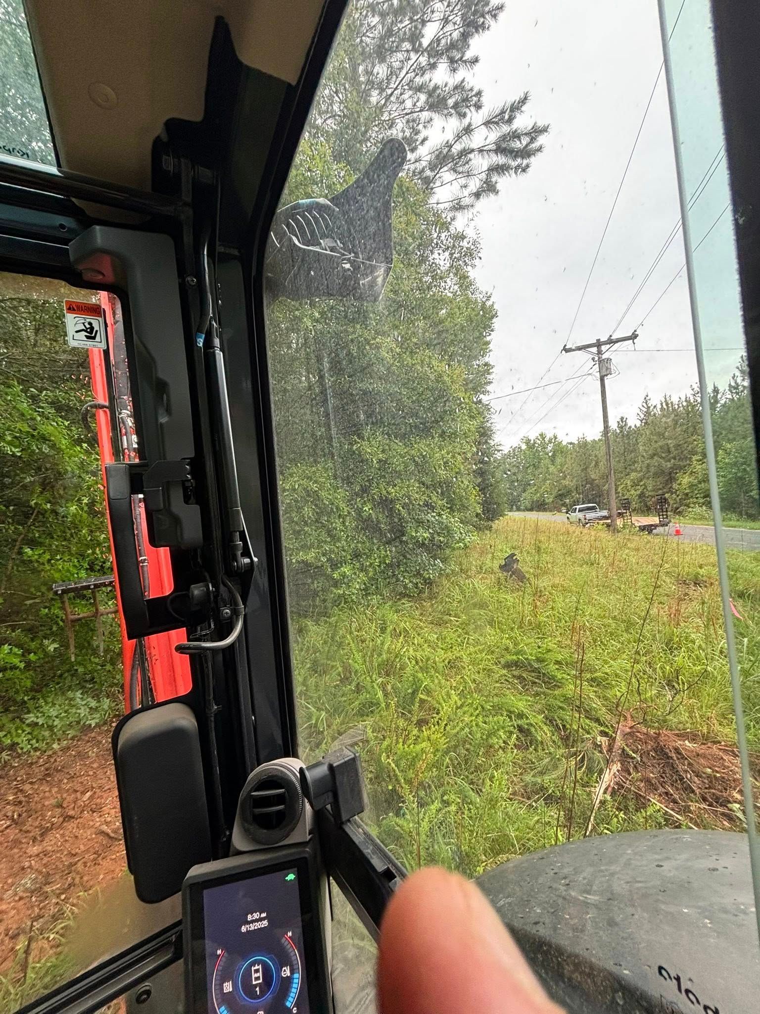 View from inside a tractor cab, showing a green landscape with a road and power lines. A finger obscures part of a screen.