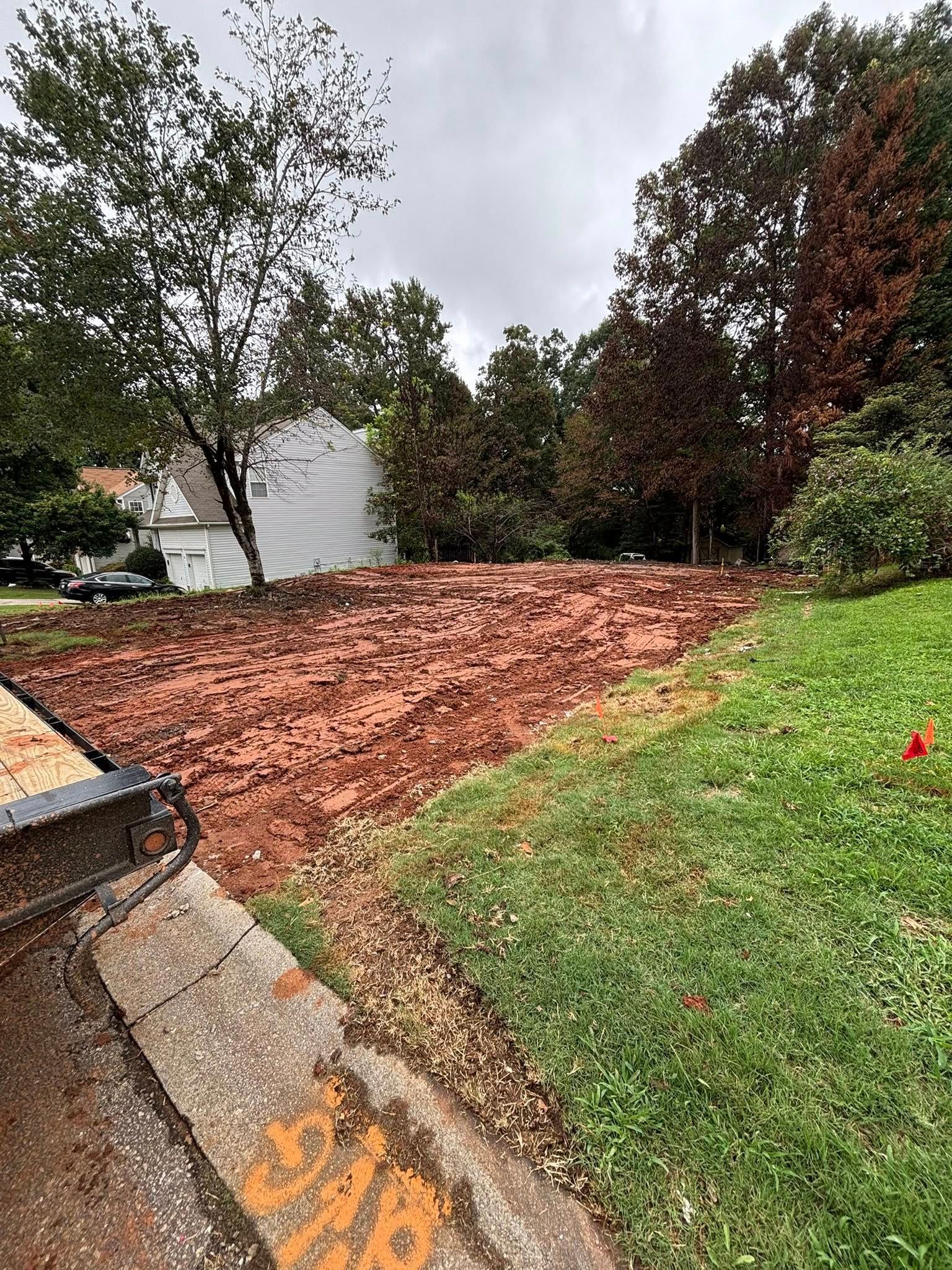 Dirt patch in a yard, bordered by grass and a sidewalk, with trees and a cloudy sky in the background.