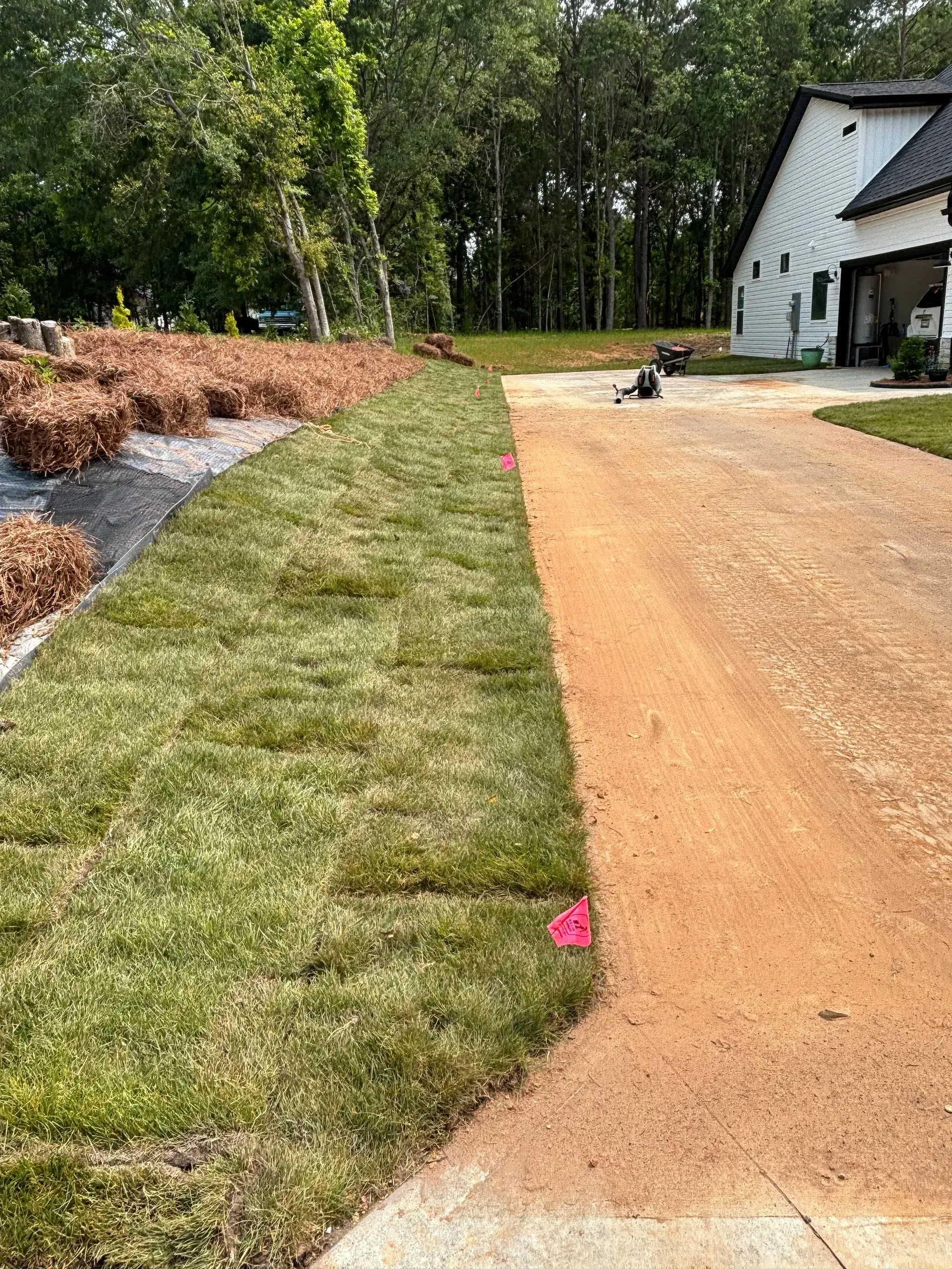 Freshly laid sod borders a gravel driveway next to a white house, a tree-lined yard is in the background.
