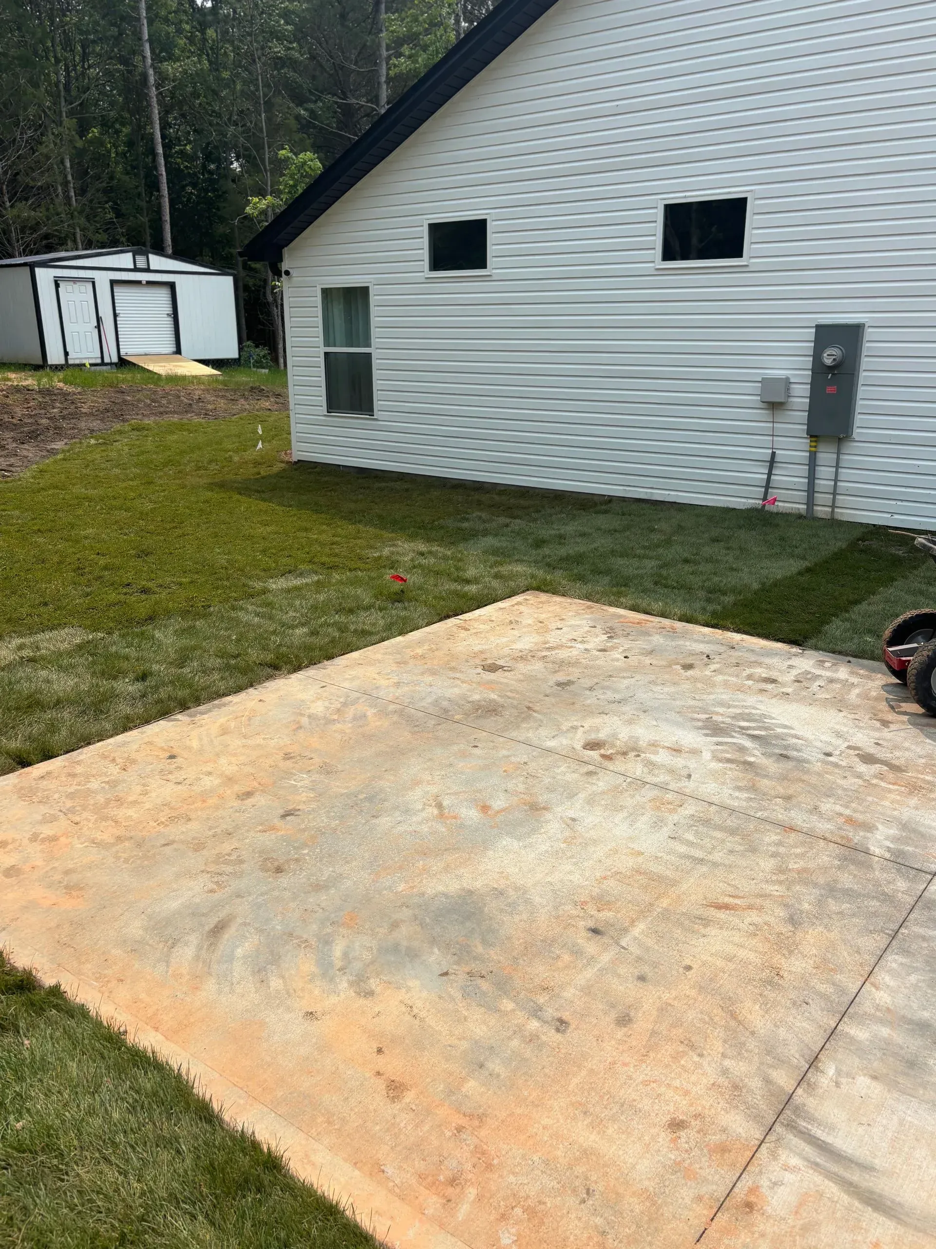 A concrete patio in front of a white-sided house with a lawn and small shed in the background.