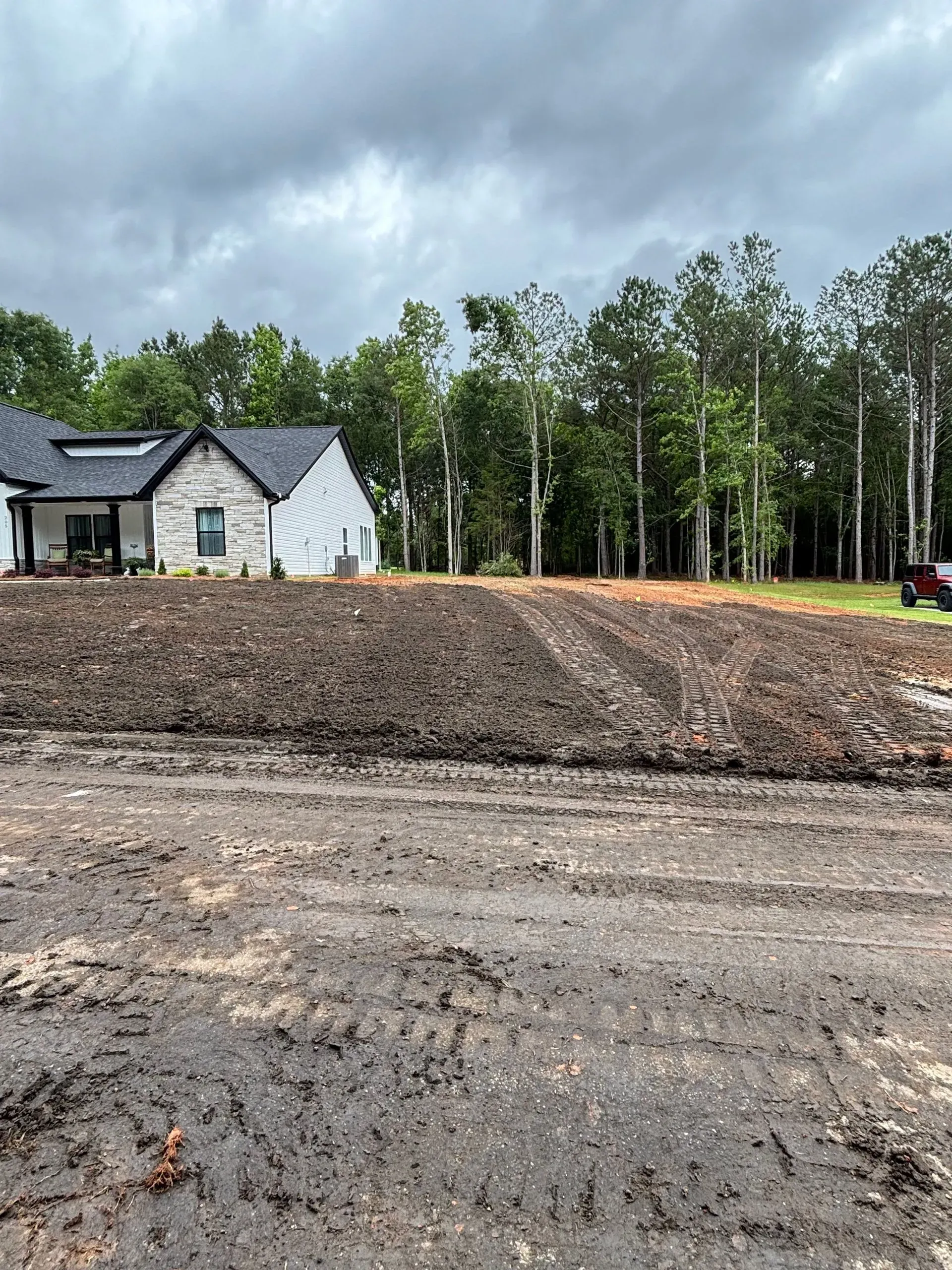 A muddy construction site in front of a modern house with a dark roof, under a cloudy sky.