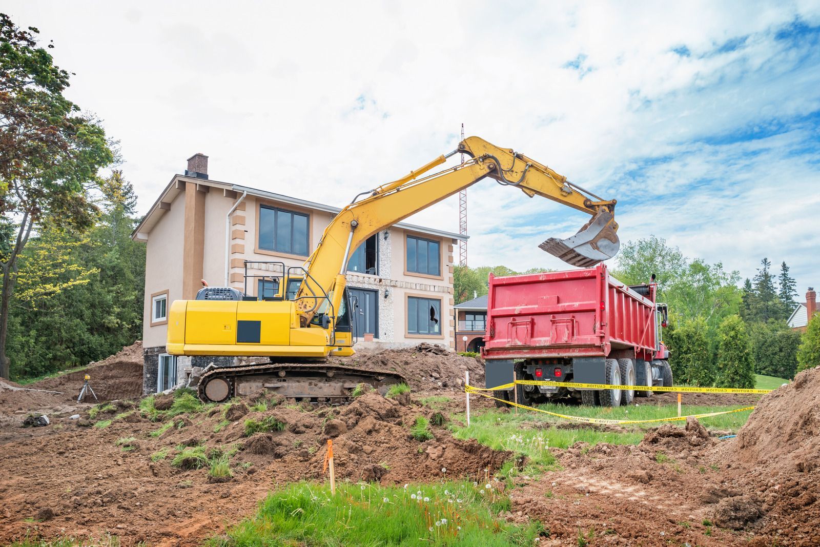 A small brick house with a chimney being demolished by an excavator; trees surround the house.