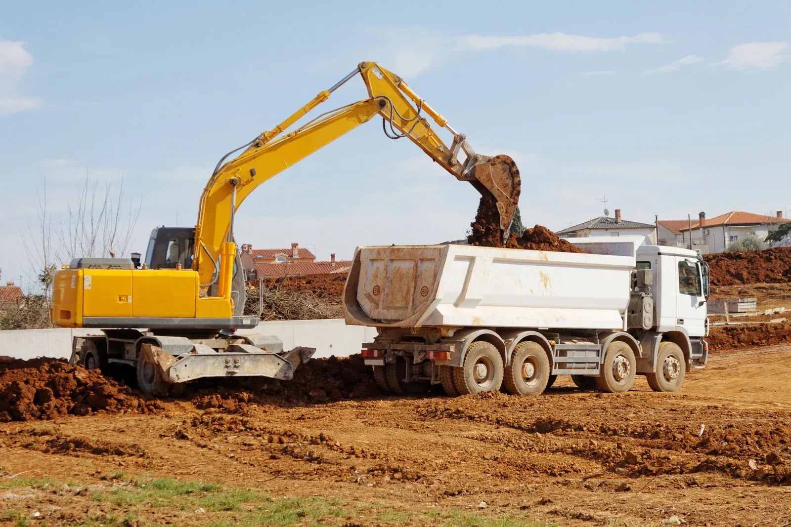 A demolished house sits among scattered debris, with a small excavator in the left foreground under a clear blue sky.