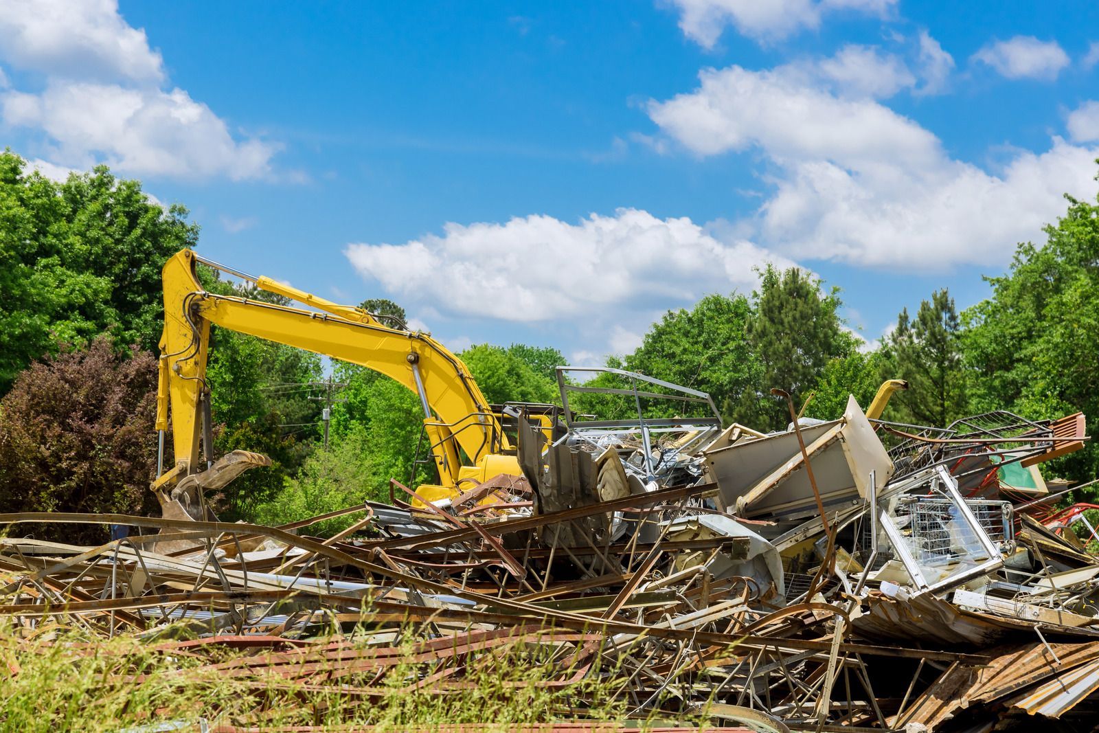 Yellow excavator demolishing building debris under a blue sky and green trees.
