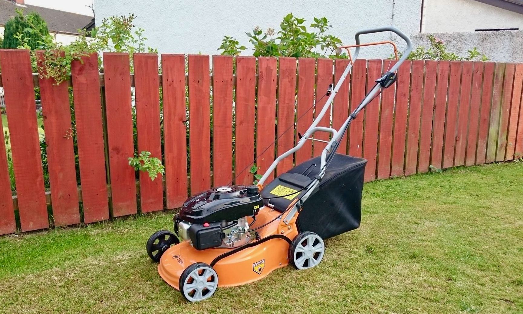 Photograph of a yellow Cyril Johnston 'Tiger' brand petrol lawnmower, parked on grass, adjacent to a red fence.
