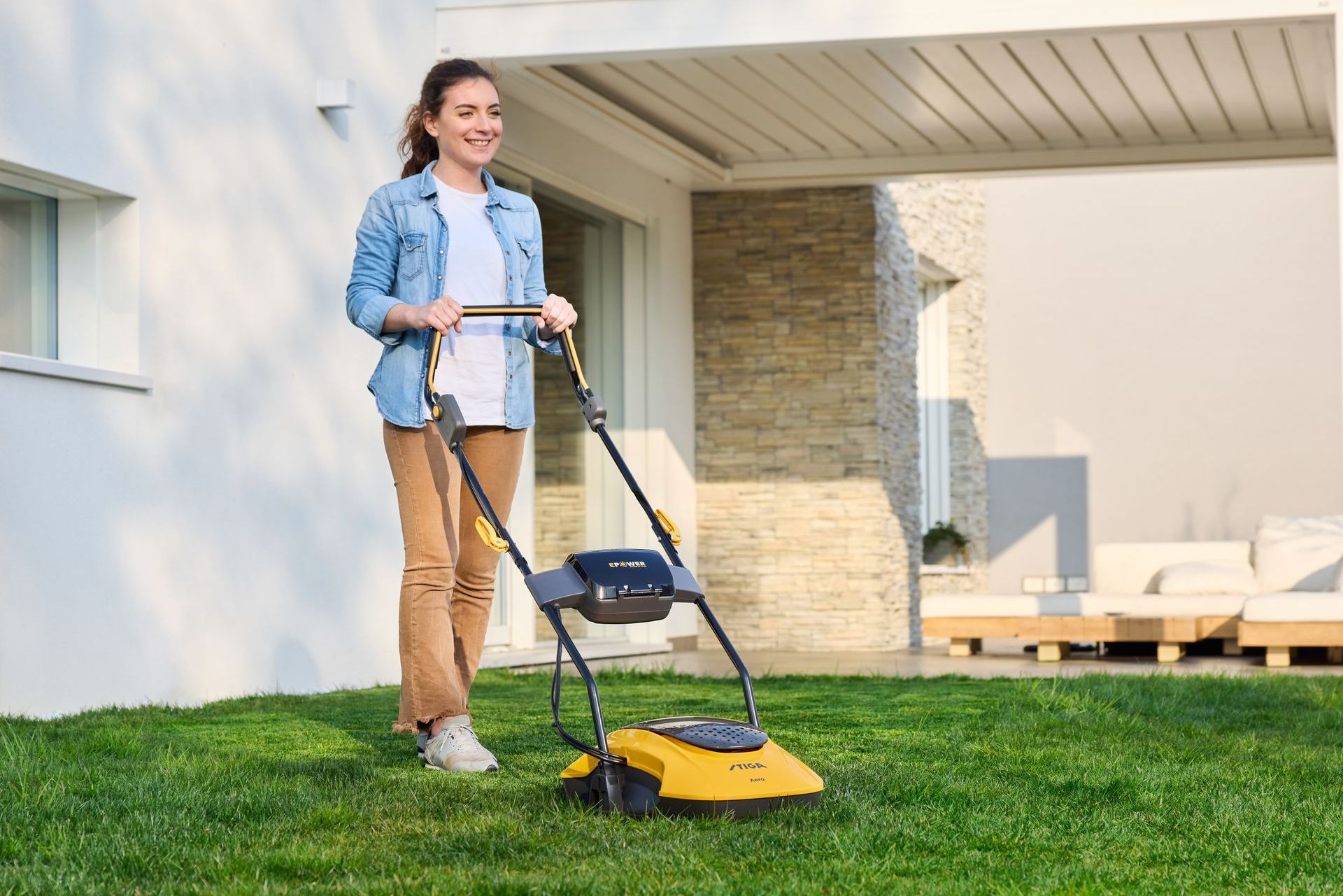 Photograph of a woman cutting grass with a yellow, STIGA brand, lithium-ion battery powered, electric hover mower.
