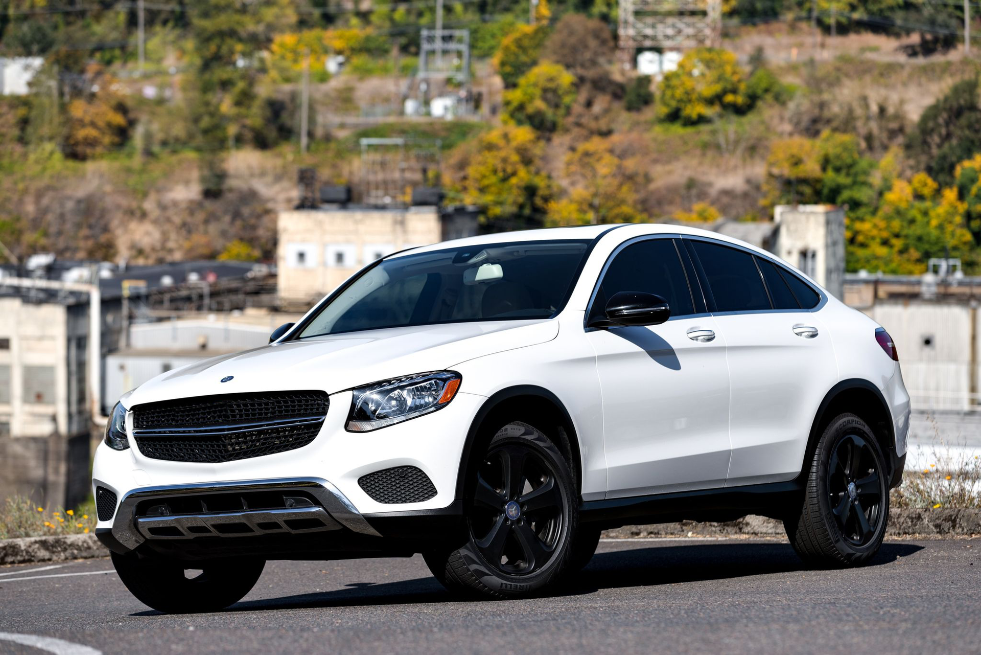 White Mercedes SUV with black wheels on a road; background includes buildings and trees.