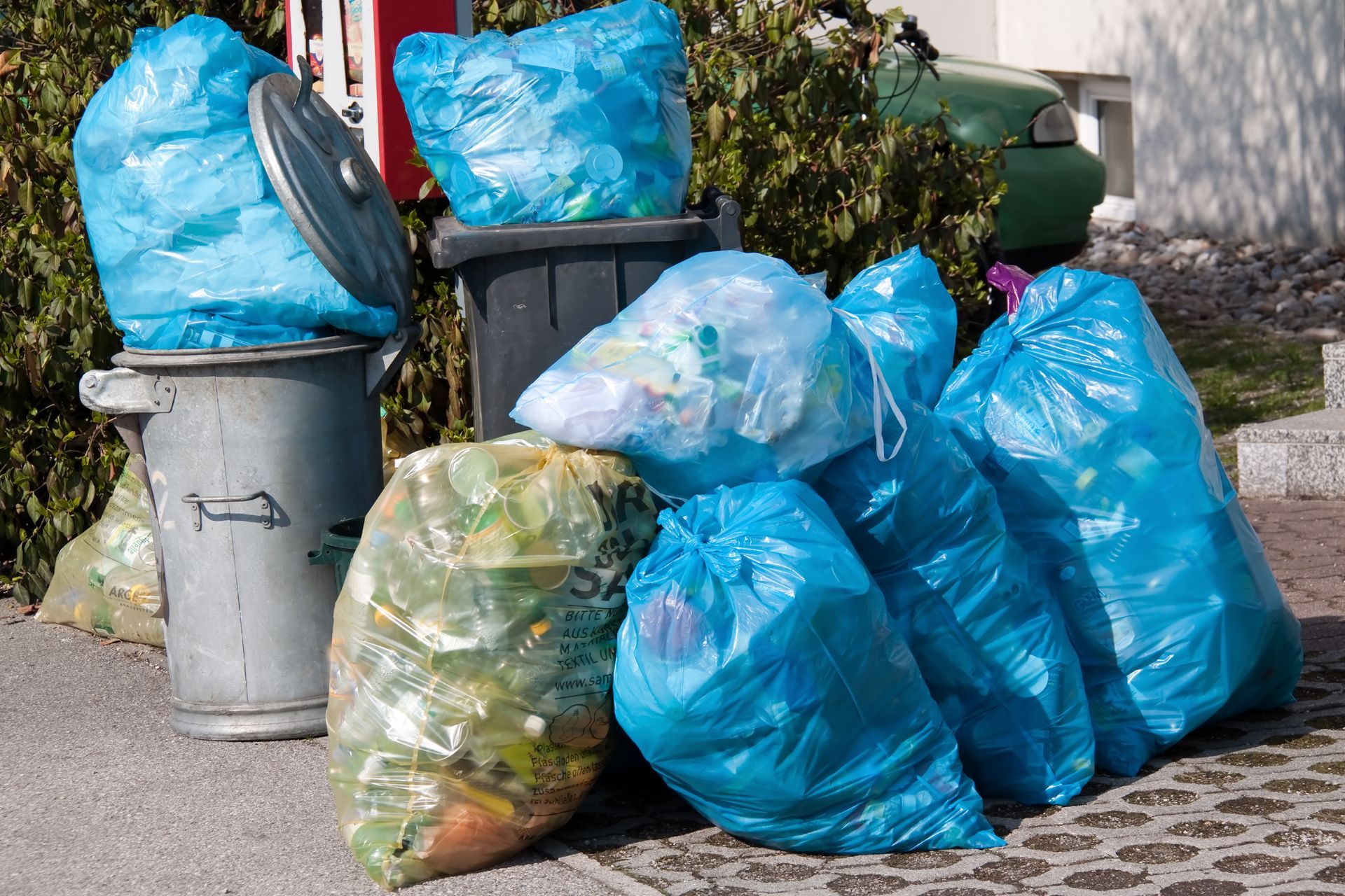 Blue and green trash bags overflowing from a trash can on a sidewalk.