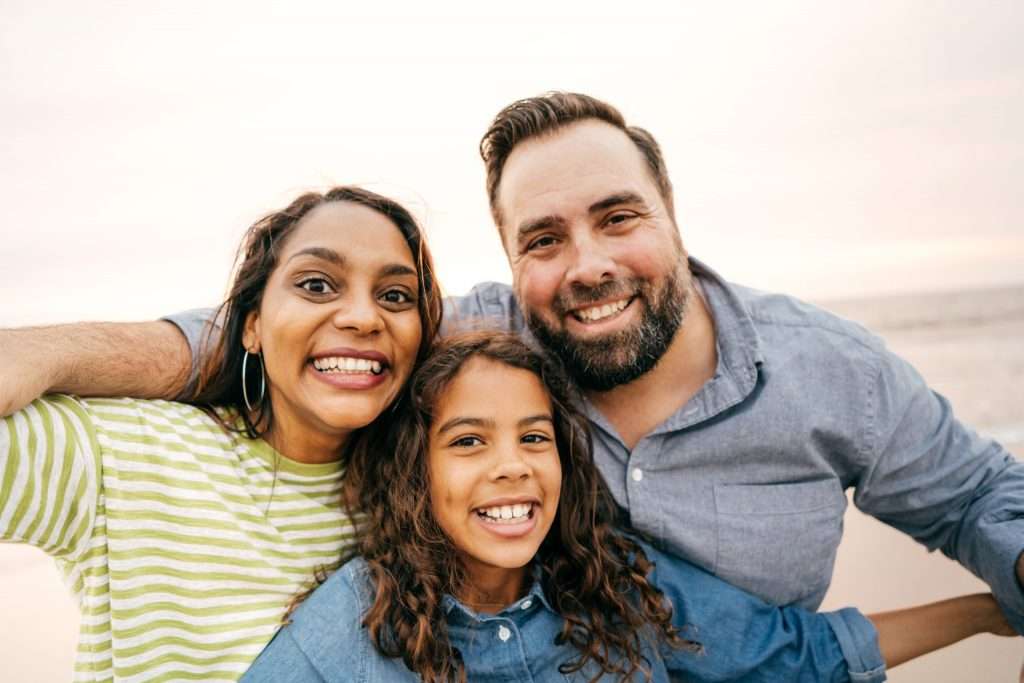 family with mom, dad and daughter standing outside by the beach smiling