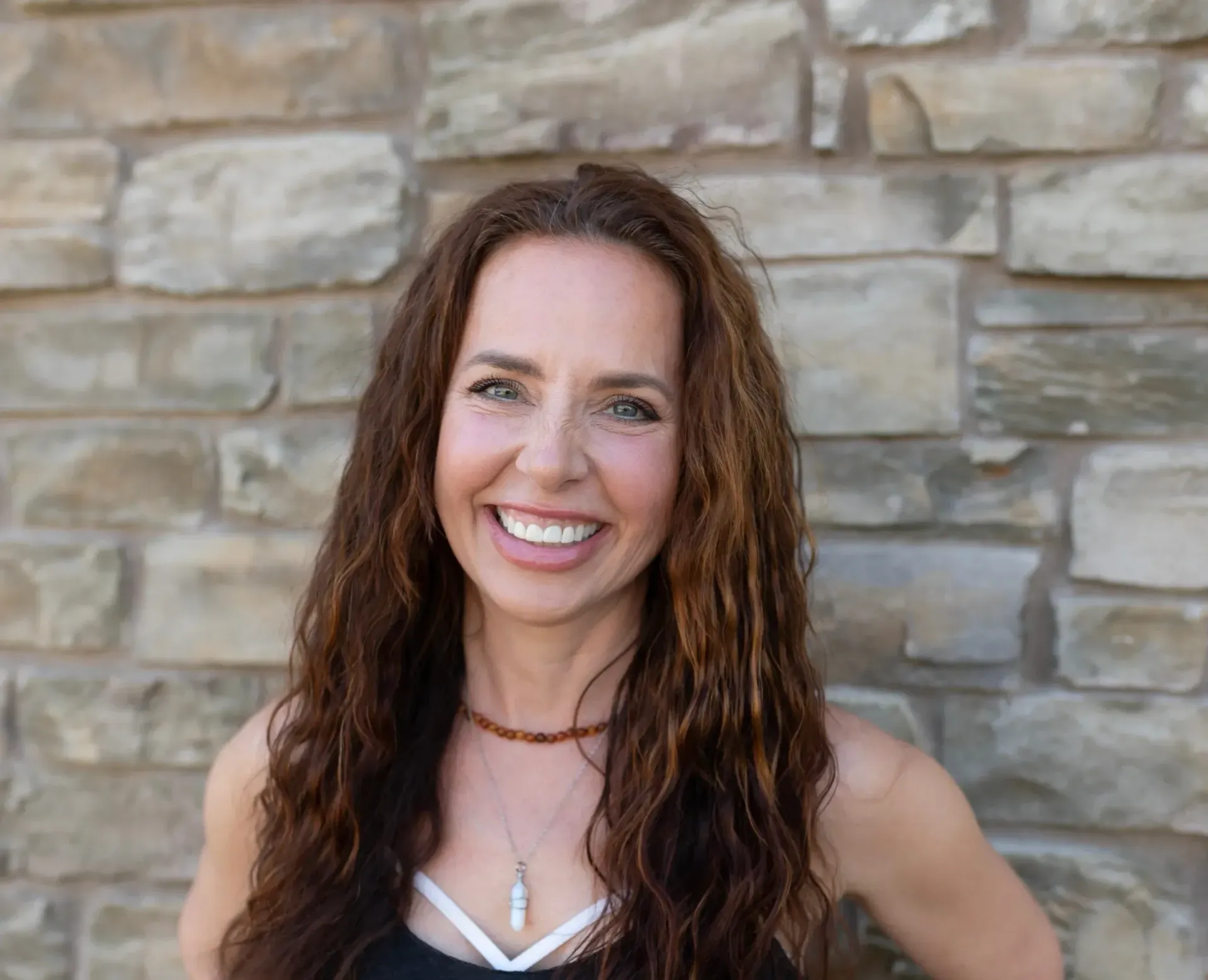 Smiling woman with long brown hair standing against a stone wall, wearing a white top and necklace