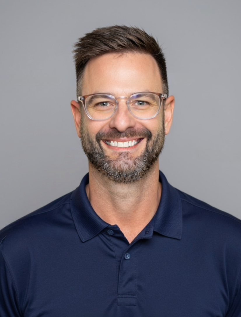 A man wearing glasses and a blue shirt is smiling in front of a white brick wall.