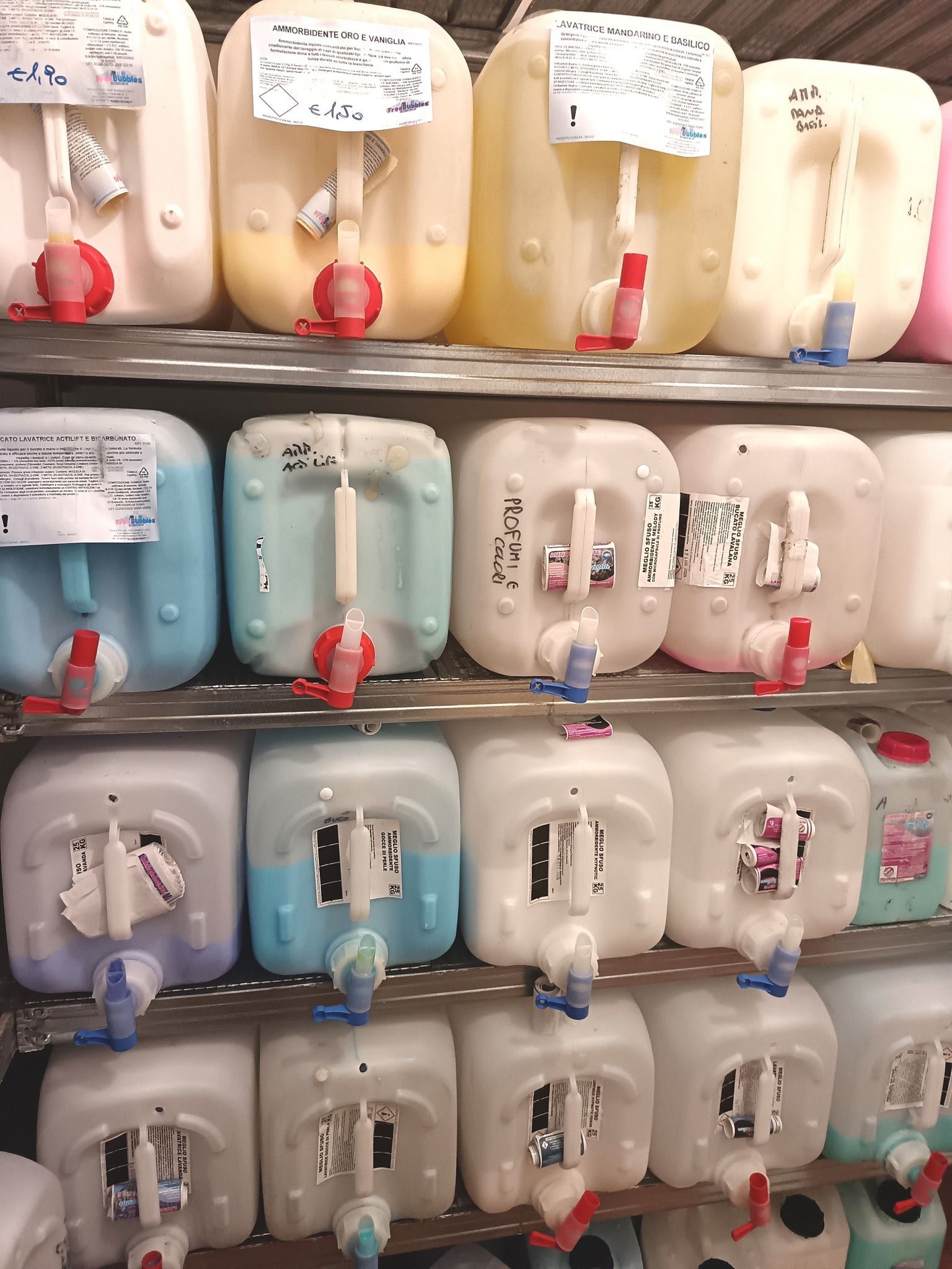 File di contenitori di plastica con rubinetti, contenenti liquidi di vari colori, etichettati sugli scaffali.